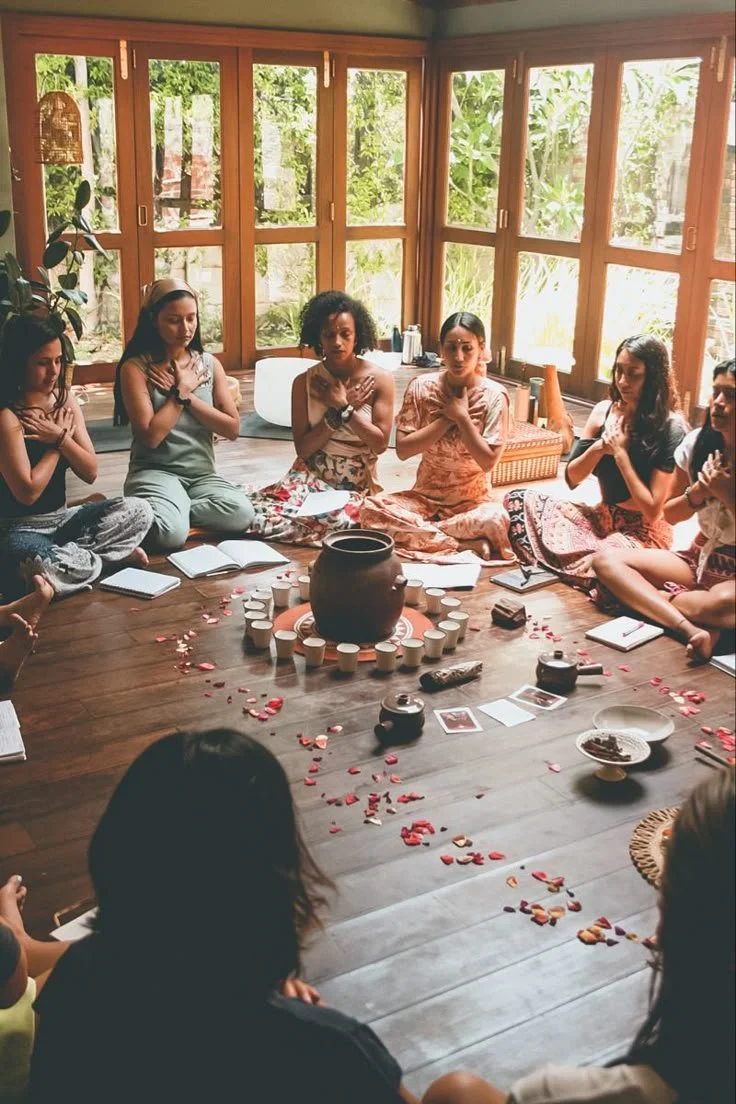 Grupo de mujeres sentadas en círculo en una madera, participando en una ceremonia o meditación, con velas, fotos y pétalos en el centro y rodeados de ventanas de cristal con vista a un jardín.