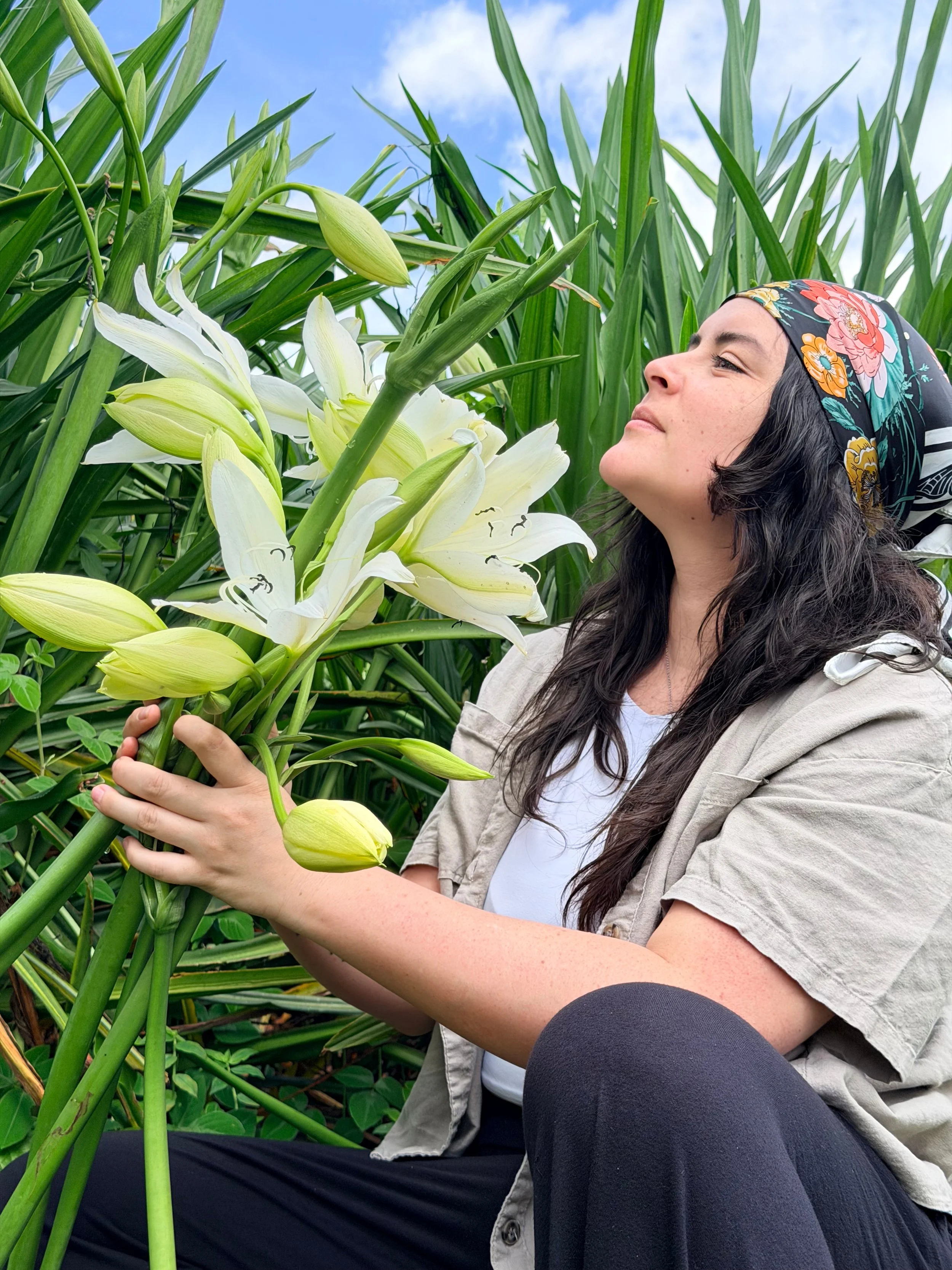 Mujer con bufanda de colores en cabeza, sonriente, sosteniendo flores de lirio blancas en un campo de plantas verdes.
