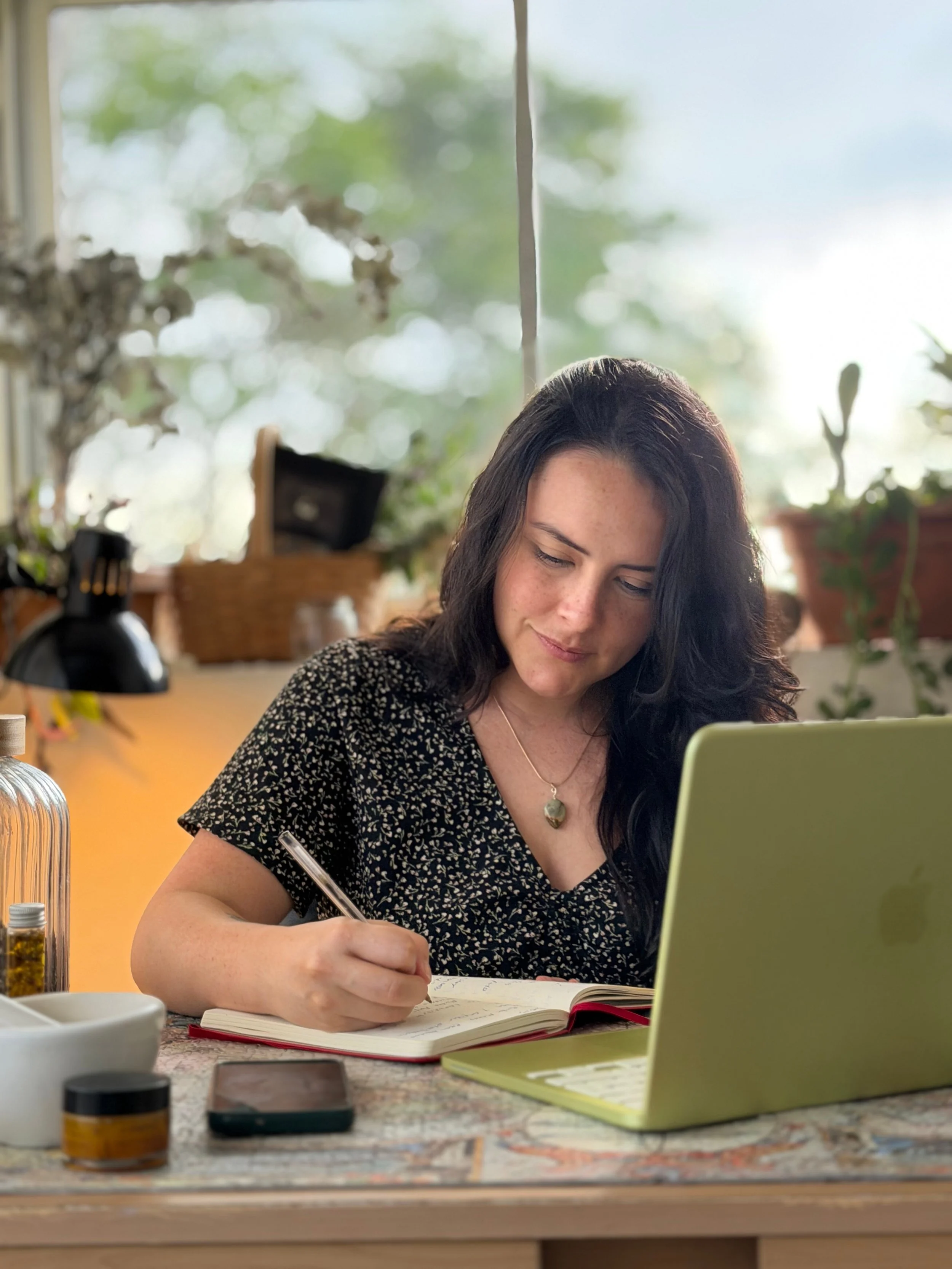 Mujer estudiando o trabajando en un escritorio con una computadora portátil, tomando notas en un cuaderno, en un ambiente con plantas y luz natural.