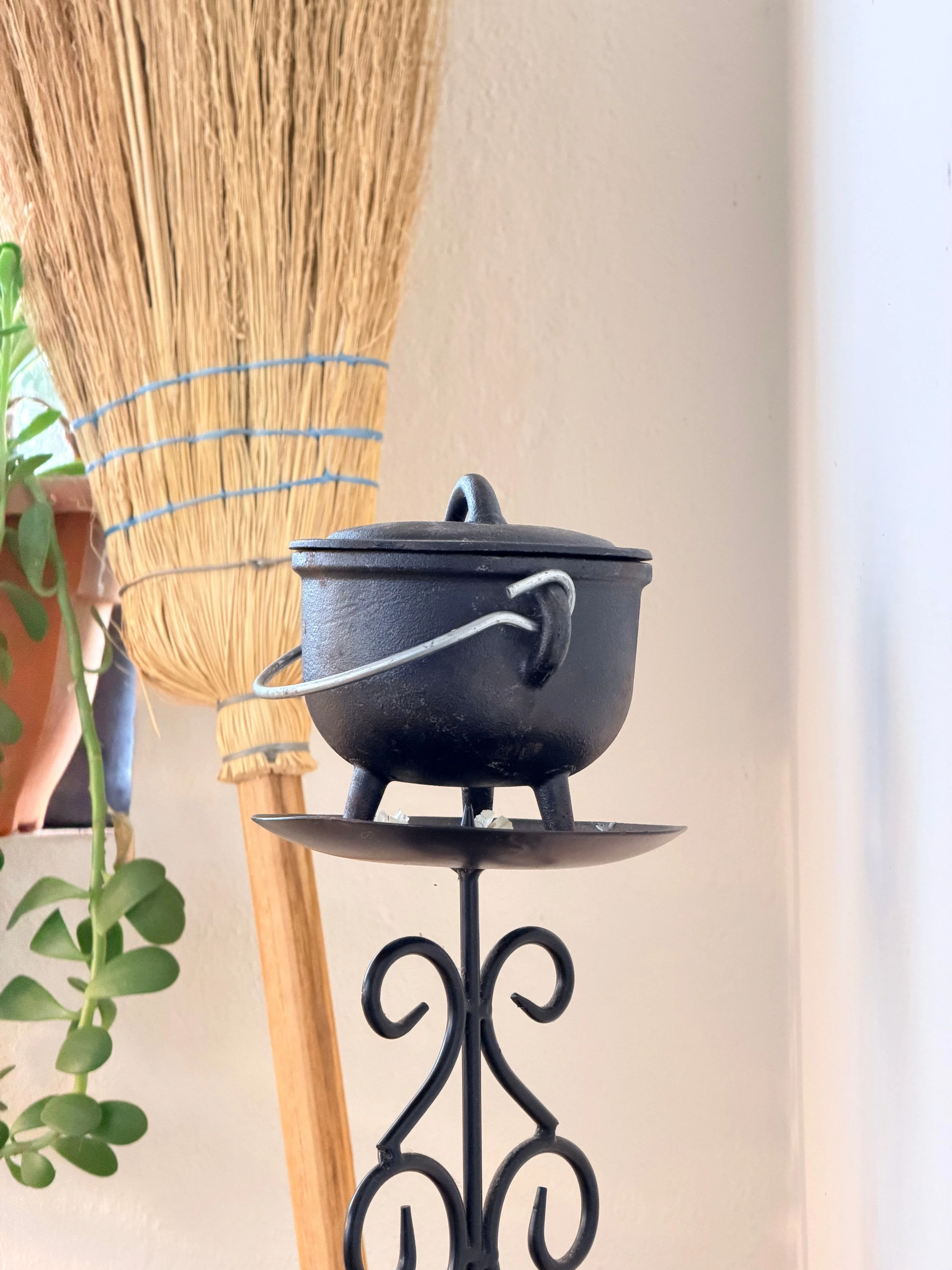 Una figura de hierro negra que sostiene una bandeja con una olla pequeña en la parte superior, en un entorno interior con pared blanca y una escoba o pala de madera a la izquierda, y una planta verde al frente.