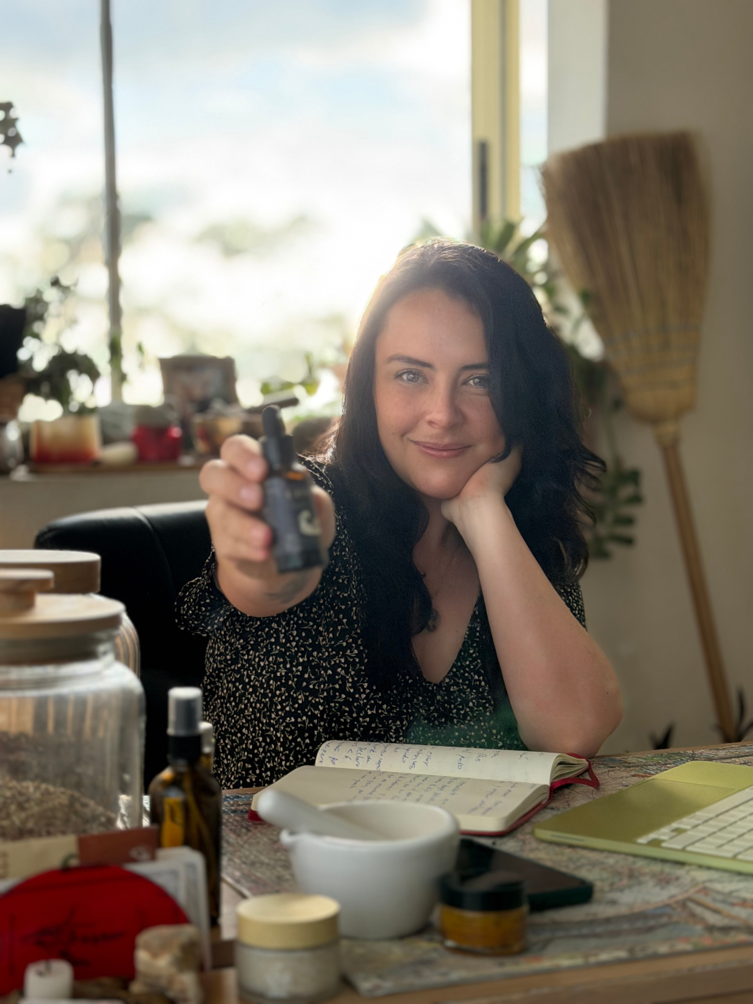 Mujer con cabello oscuro y ojos claros, sonriendo, sosteniendo un gotero en una mano en un ambiente interior con luz natural y plantas de fondo. Frente a ella hay varios objetos, incluyendo libros y frascos.