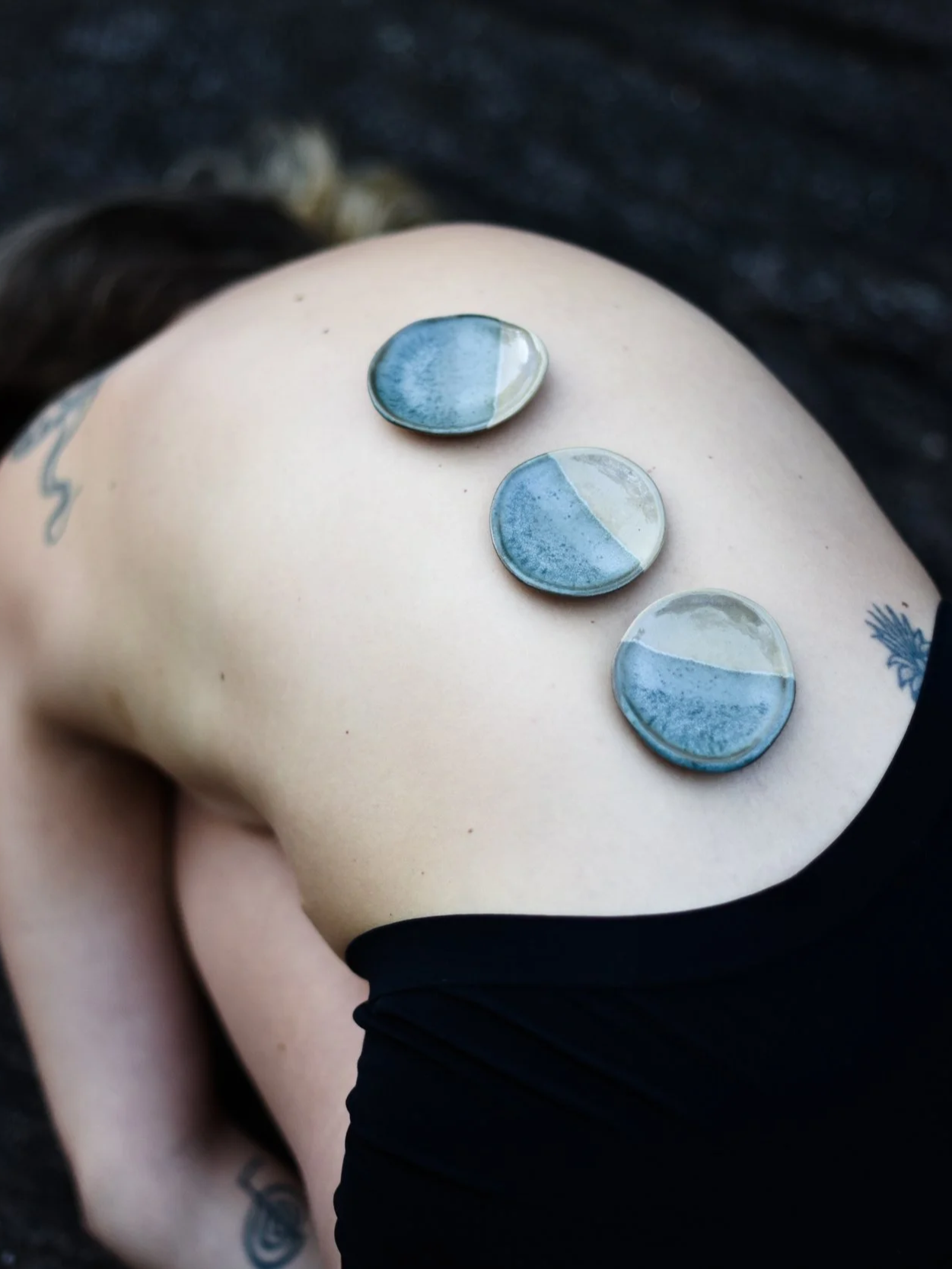 Close-up of a person's side torso with three small circular stones balanced on the skin, showing a two-tone blue and beige pattern, with a black background.