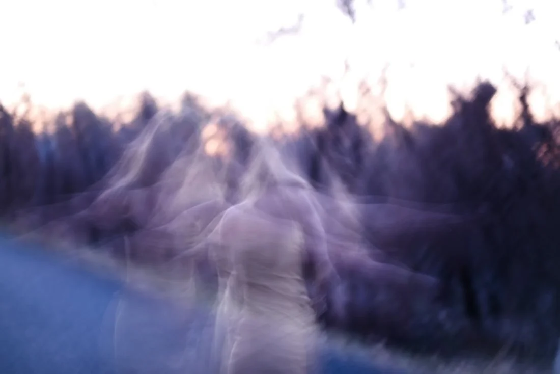 A ghostly, blurry figure appearing to walk on an outdoor path during sunset with trees in the background.