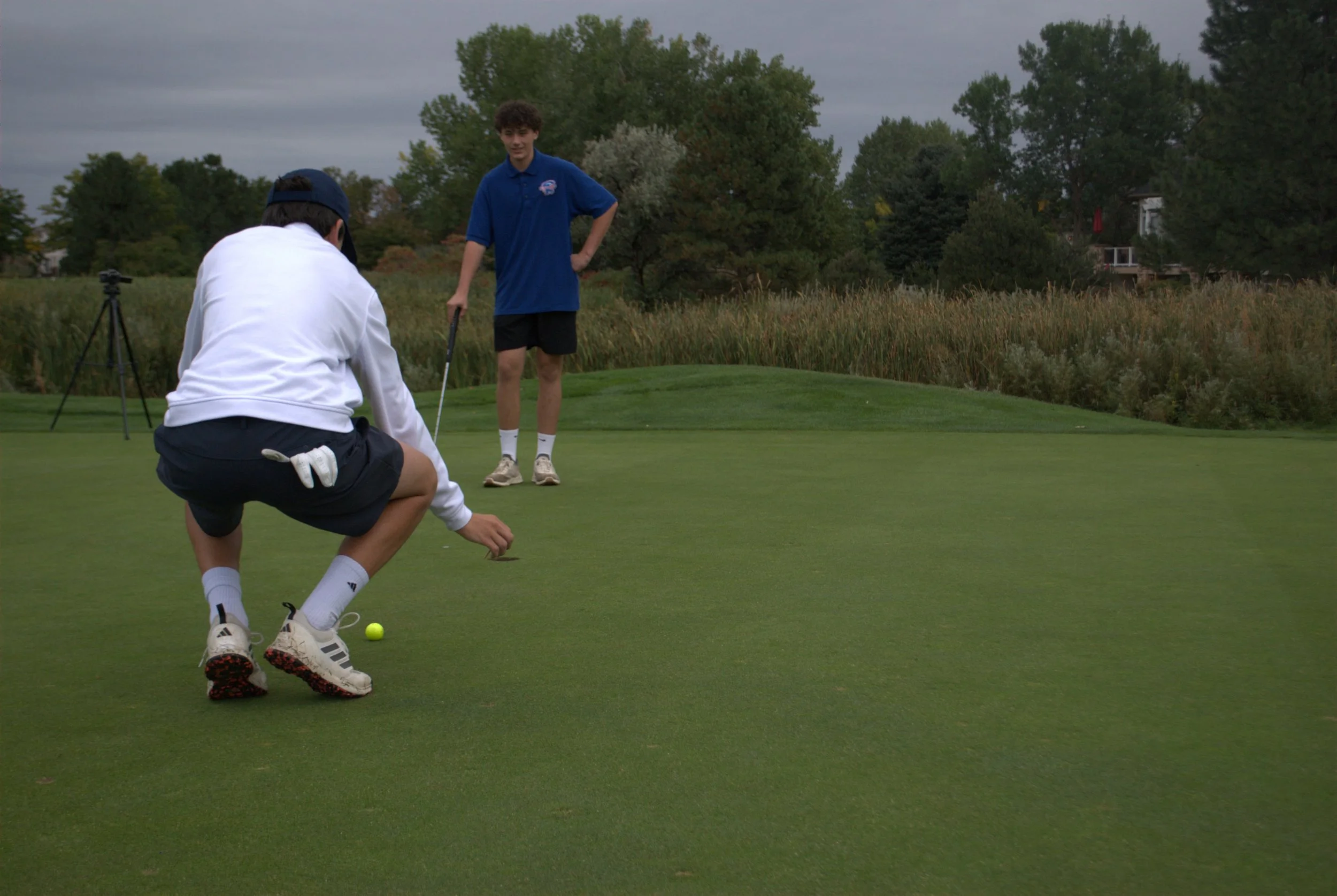 Content creation with 18 Fours Media. Two boys on a golf course, one preparing to putt and the other watching, with trees and cloudy sky in the background during daytime.