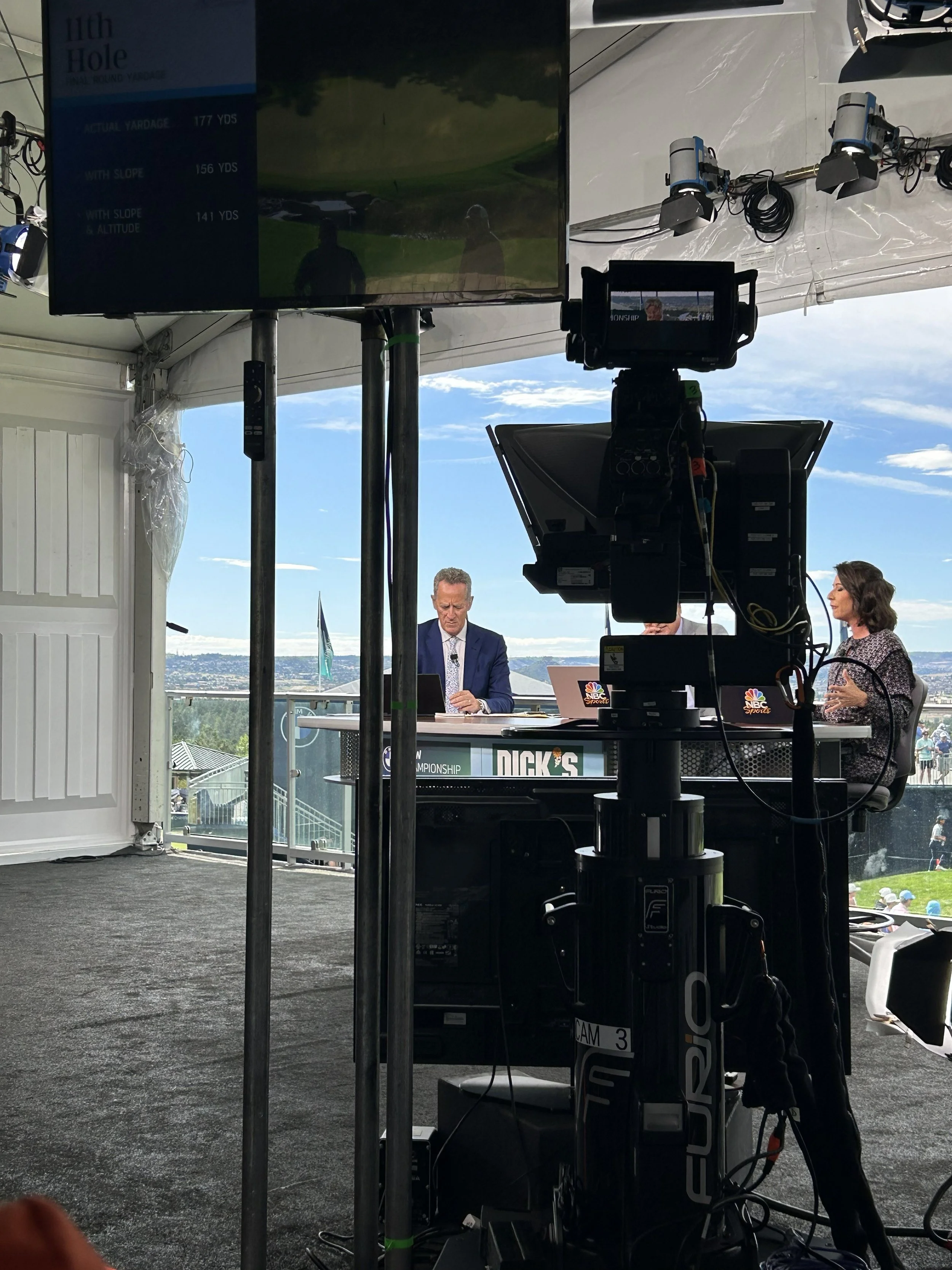 Golf Channel behind the scenes. Television screen displaying golf information, set up inside a temporary event tent with people visible outside on a sunny day. Camera equipment positioned in front and to the side of the television.