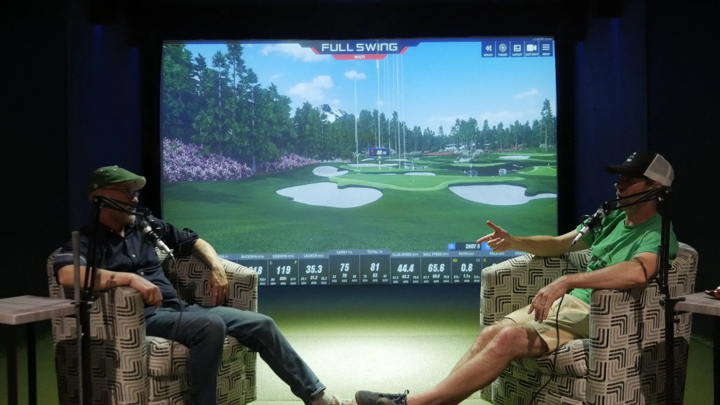 Two people sitting in chairs indoors, facing a large screen displaying a golf simulation image of a golf course, with various data overlays and statistics.