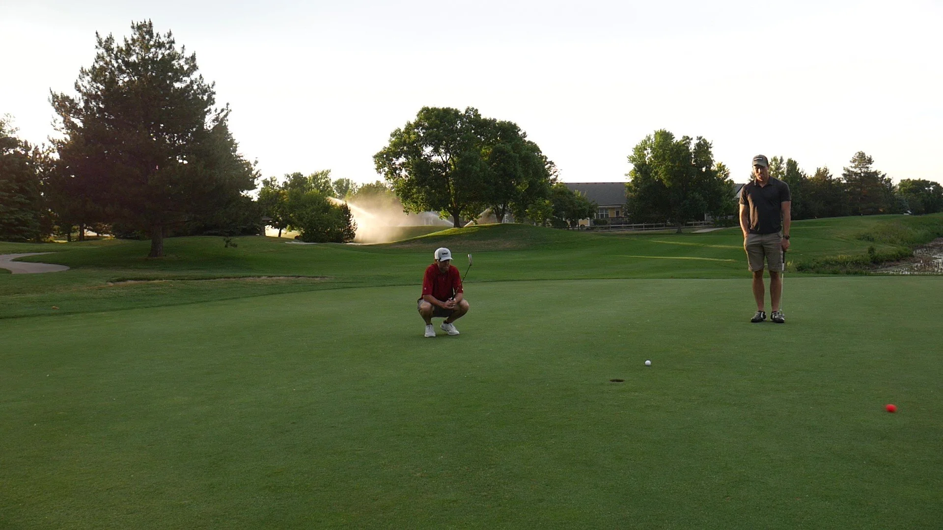Two men on a golf course preparing to putt the ball, with trees and a building in the background, during late afternoon or early evening.