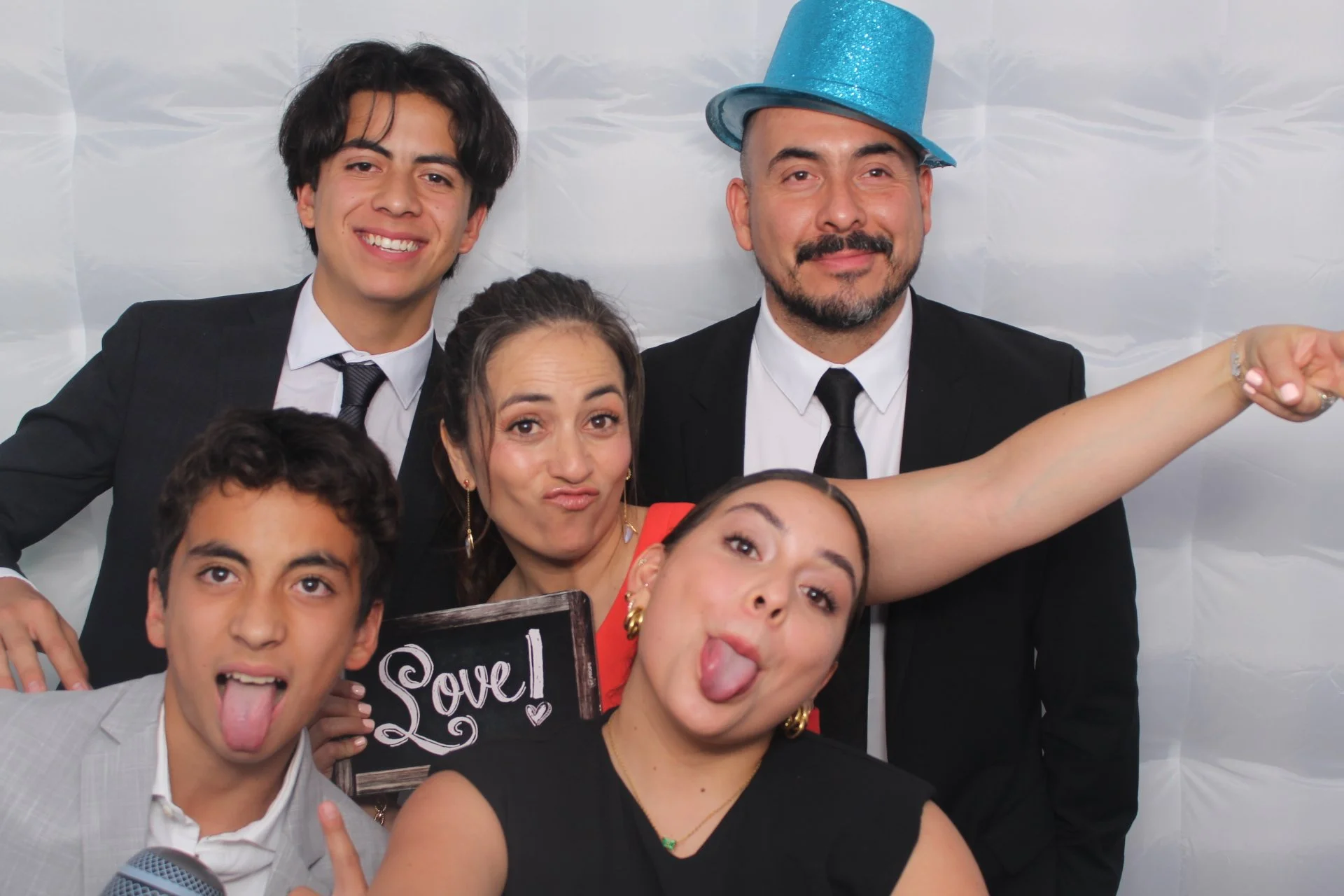 Group of people at a celebration, with some sticking out tongues and one person wearing a blue glittery top hat, against a white backdrop.