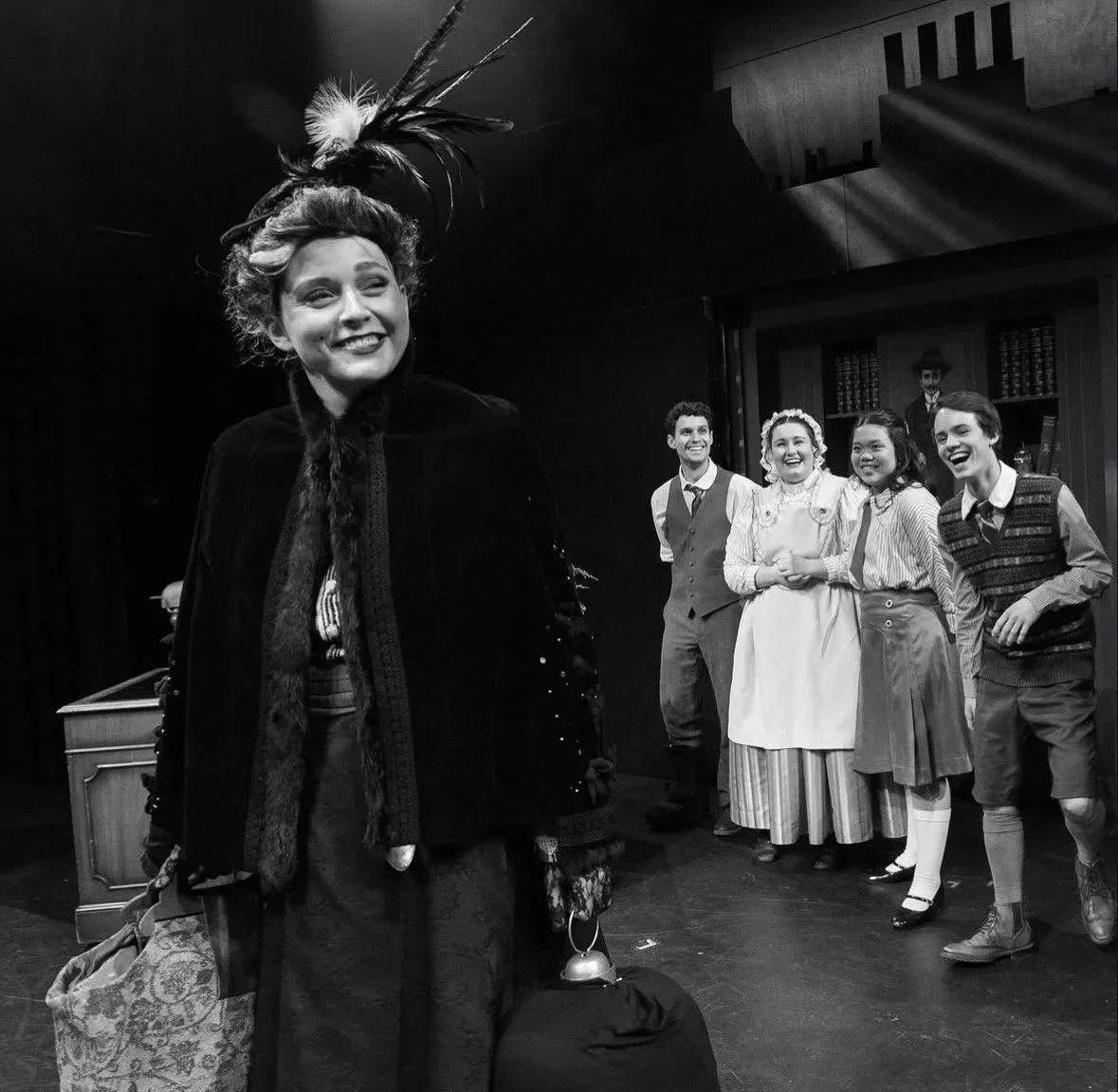 A woman dressed in vintage costume with a feathered hat smiling and holding a small bell, on stage with four other actors in period clothing, in a theatrical performance setting.