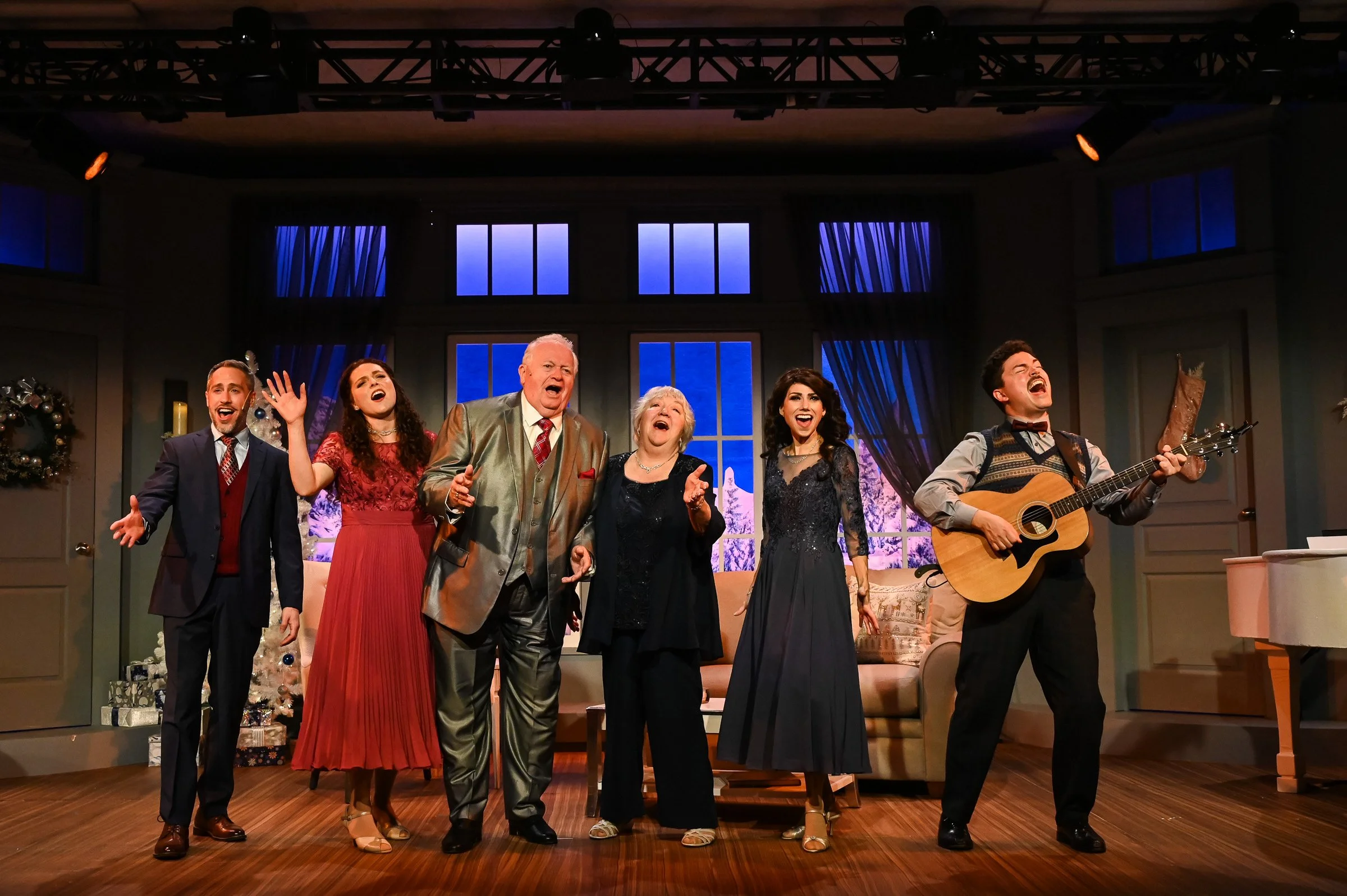 A group of six people performing on a stage, singing and playing guitar, in a cozy room with Christmas decorations, including a small Christmas tree and wrapped gifts, during a holiday gathering.