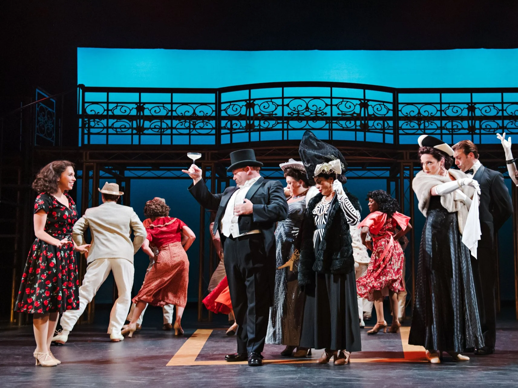 The image depicts a lively stage scene with actors dressed in vintage clothing, possibly from the 1950s or 1960s. The scene includes a woman in a floral dress, men in suits and hats, and women in elegant dresses with large hats, engaged in a dance or theatrical performance. The background features a dark set with a blue-lit section and an ornate balcony structure.