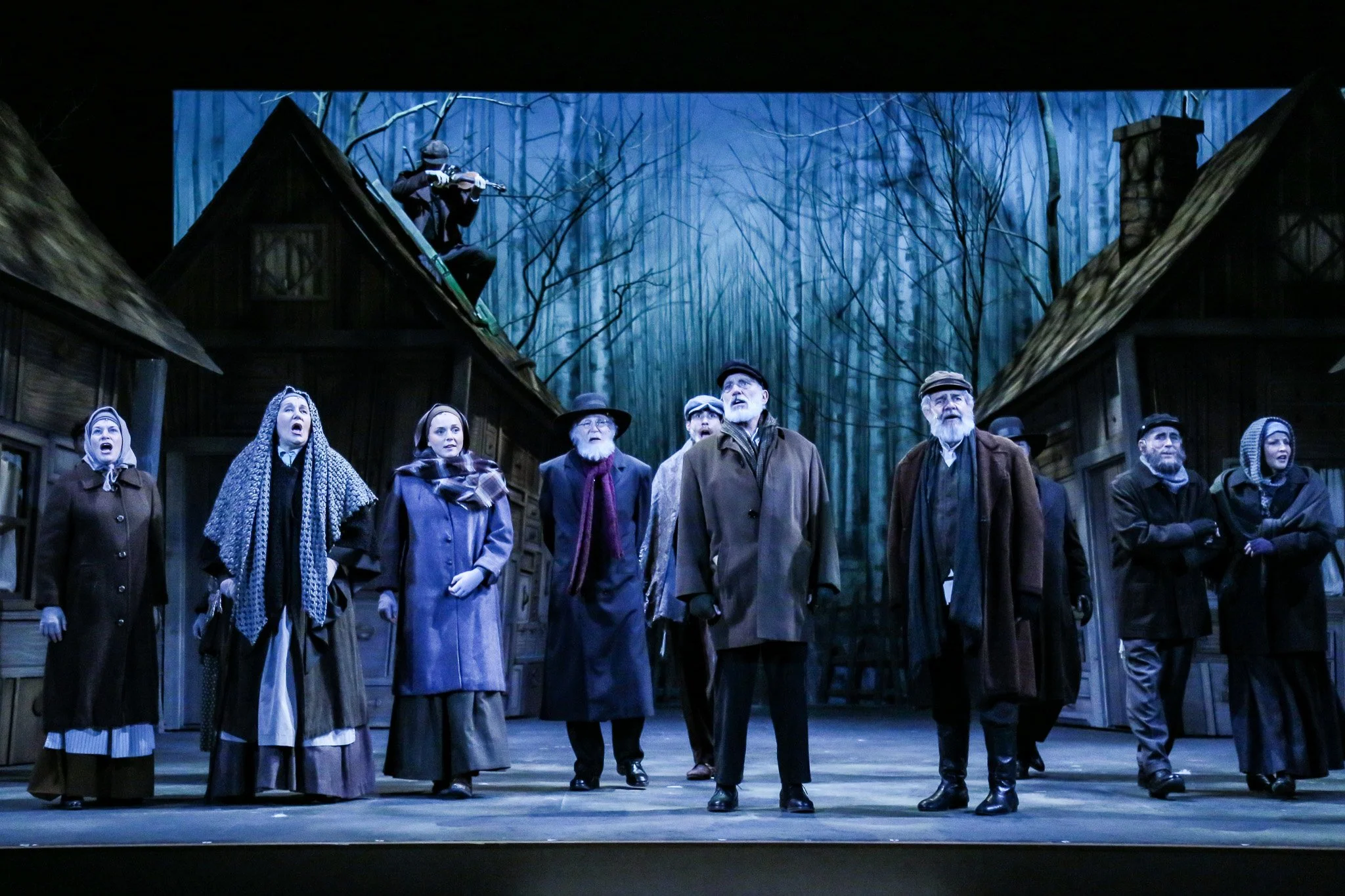 A theatrical scene with a group of people dressed in 19th-century clothing, standing outdoors in a wooded area, with a man on the roof aiming a rifle, set against a background of tall trees and wooden buildings.