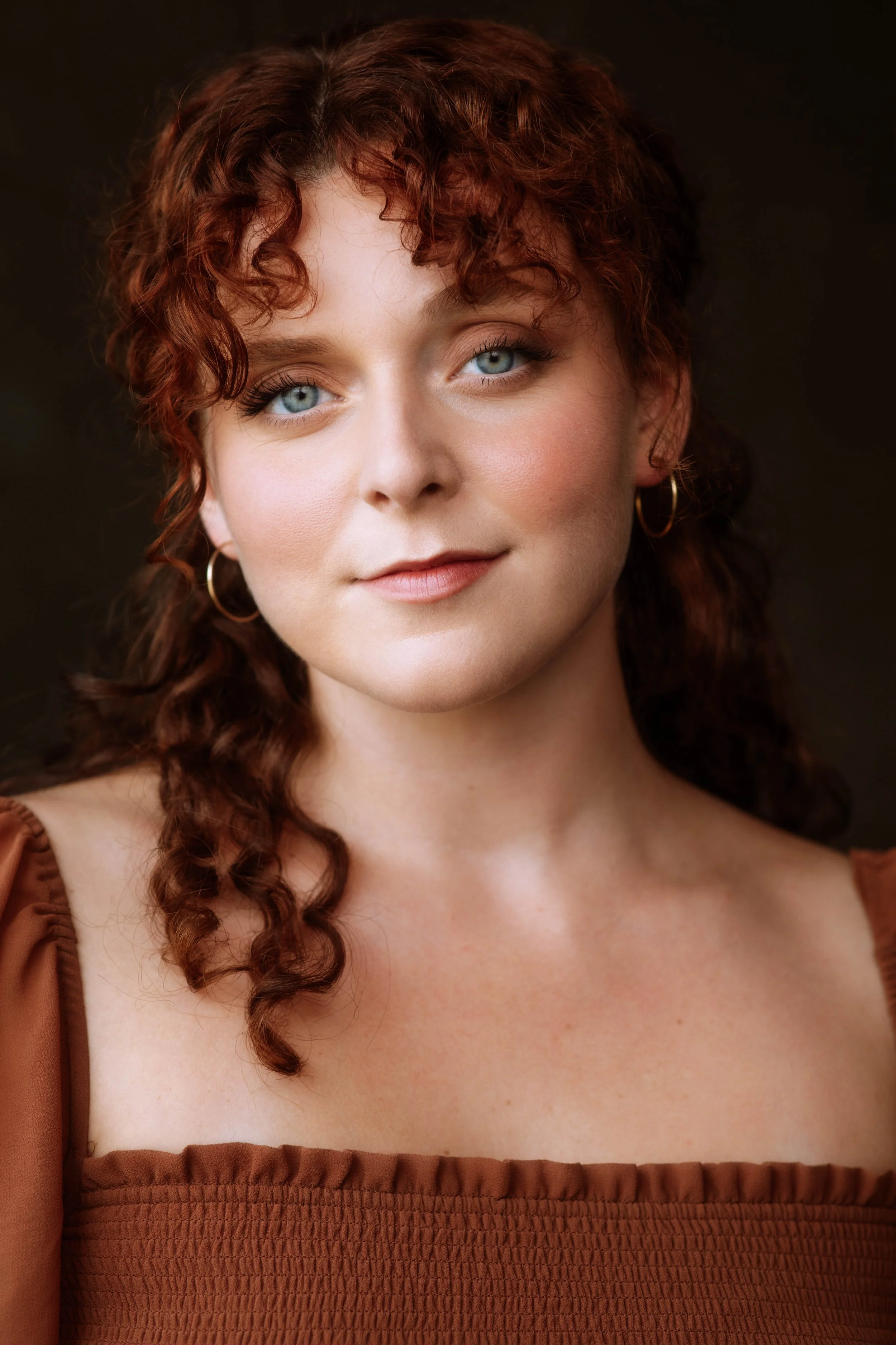 A woman with curly red hair and blue eyes posing against a dark background, wearing a brown sleeveless top with gathered fabric.