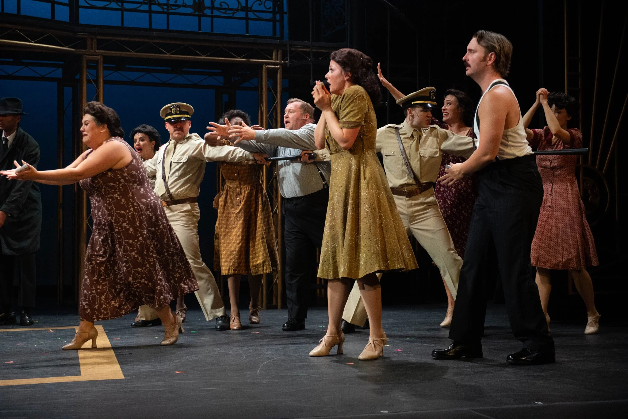 Stage scene from a musical with performers in vintage costumes, including military uniforms and elegant dresses, dancing and singing under stage lighting.