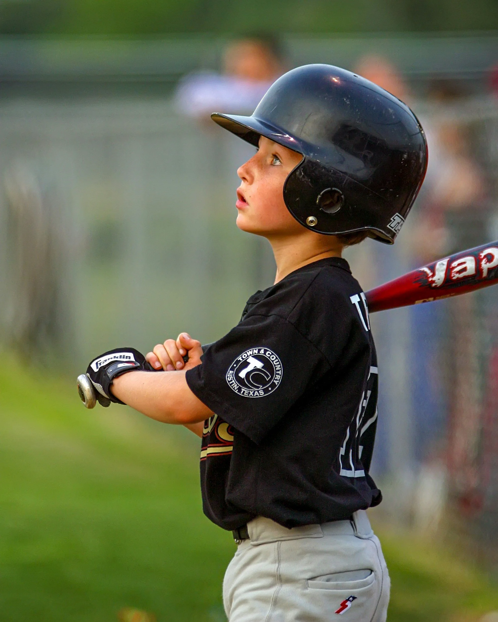 Young baseball player with a black helmet and black shirt, holding a red baseball bat, standing outdoors on a baseball field.