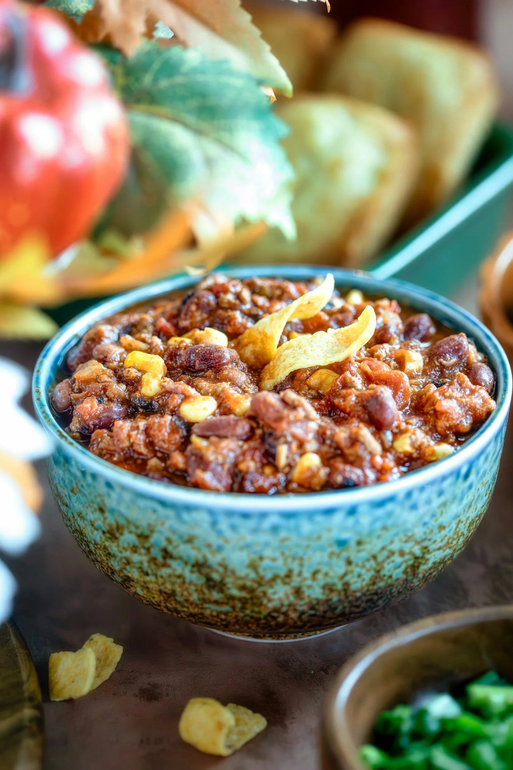 Close-up of a bowl of chili with beans and minced meat, garnished with cheese chips, surrounded by other ingredients including tomatoes, cheese, and green vegetables.