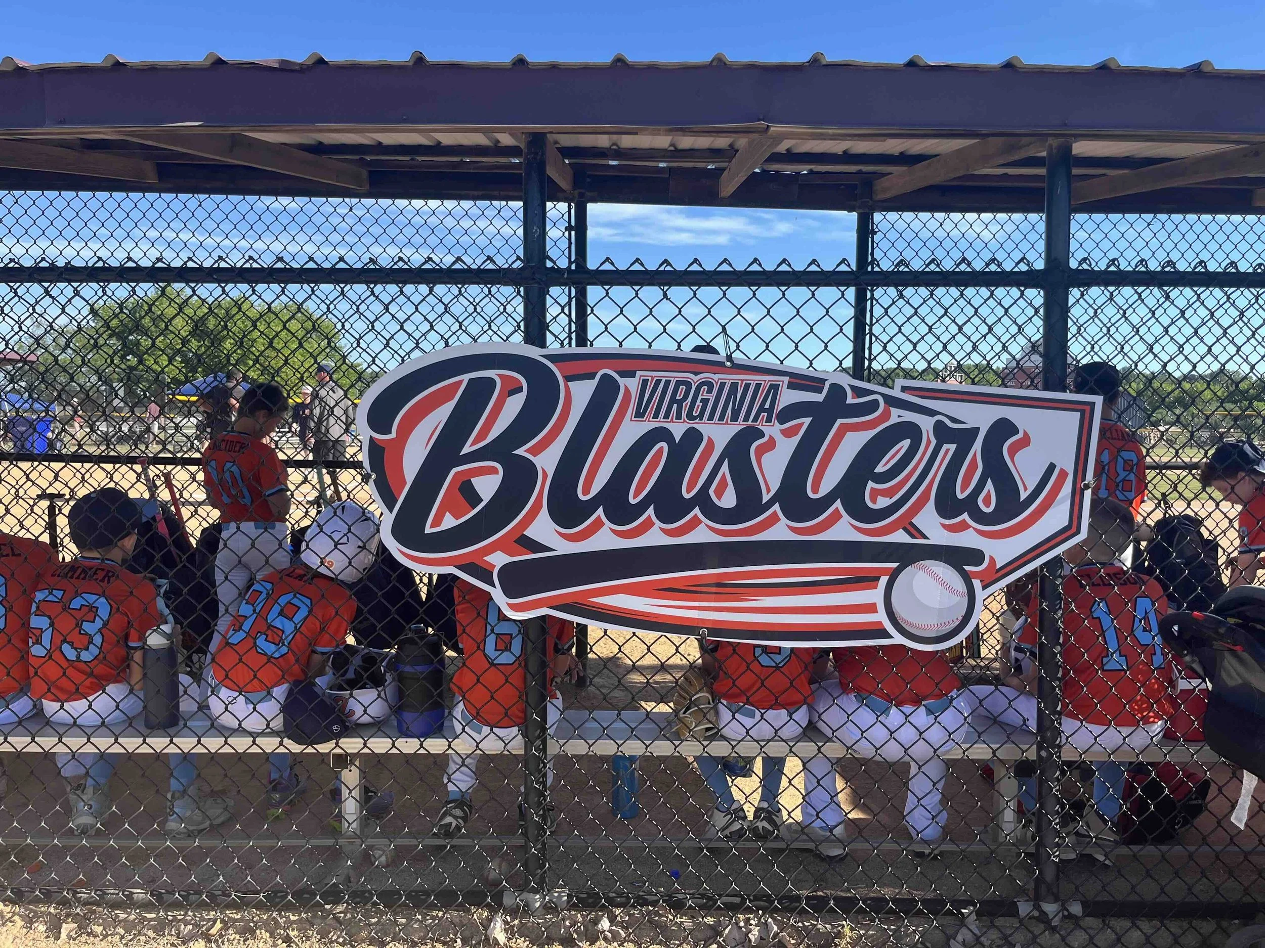 Baseball players in orange and blue uniforms sitting on a bench behind a chain-link fence at a baseball game. A sign that reads "Virginia Blasters" is hanging on the fence.