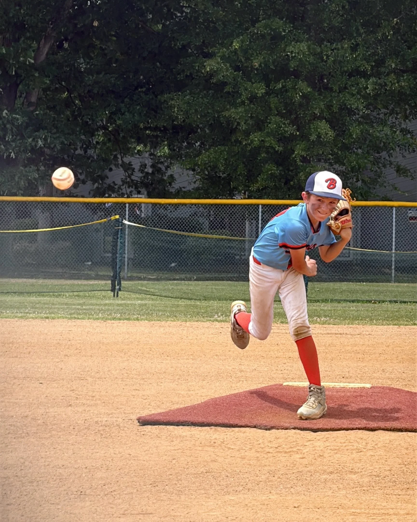 Young baseball player in a blue uniform and cap, smiling, stands on a mound in a baseball field, preparing to pitch the ball.