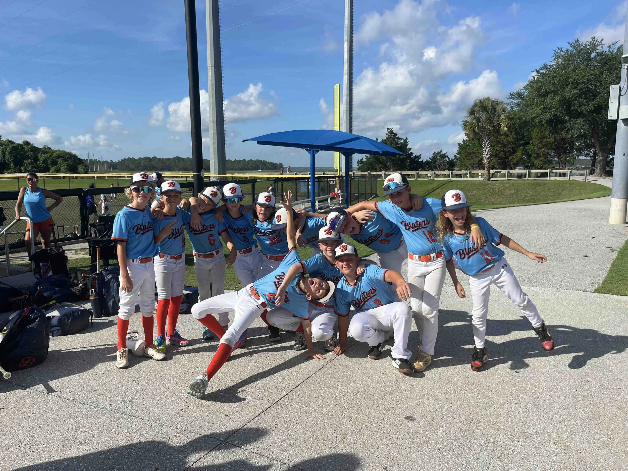 A group of young baseball players in blue jerseys and white pants posing together, some making silly faces and poses, under a bright blue sky with scattered clouds, near a baseball field with a fence and trees in the background.