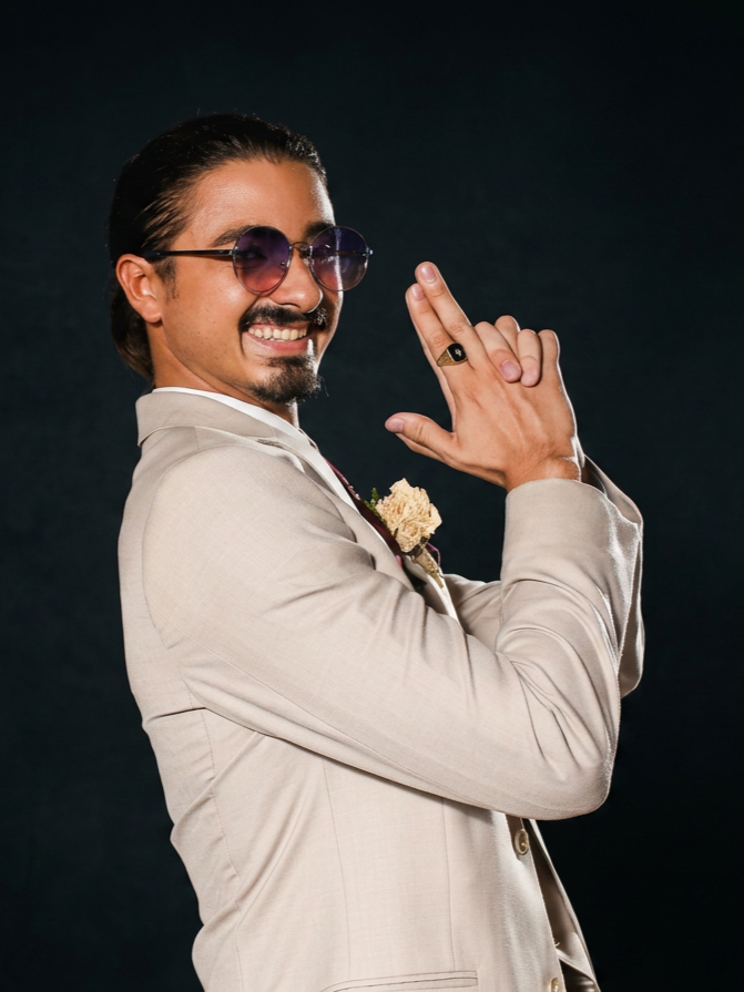 Man with dark hair, mustache, and beard wearing purple-tinted sunglasses and a beige suit, smiling and posing with hands clasped together, against a dark background. Author