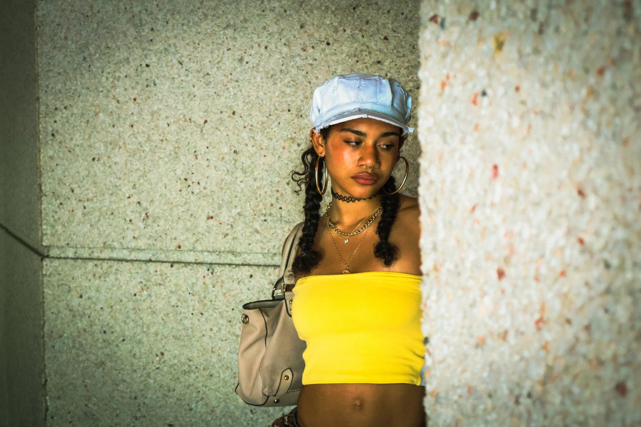 A young woman with braided hair, hoop earrings, and layered necklaces leaning against a speckled concrete wall. She is wearing a yellow tube top and has a beige backpack hanging from her shoulder. She has a neutral expression and appears to be lookin