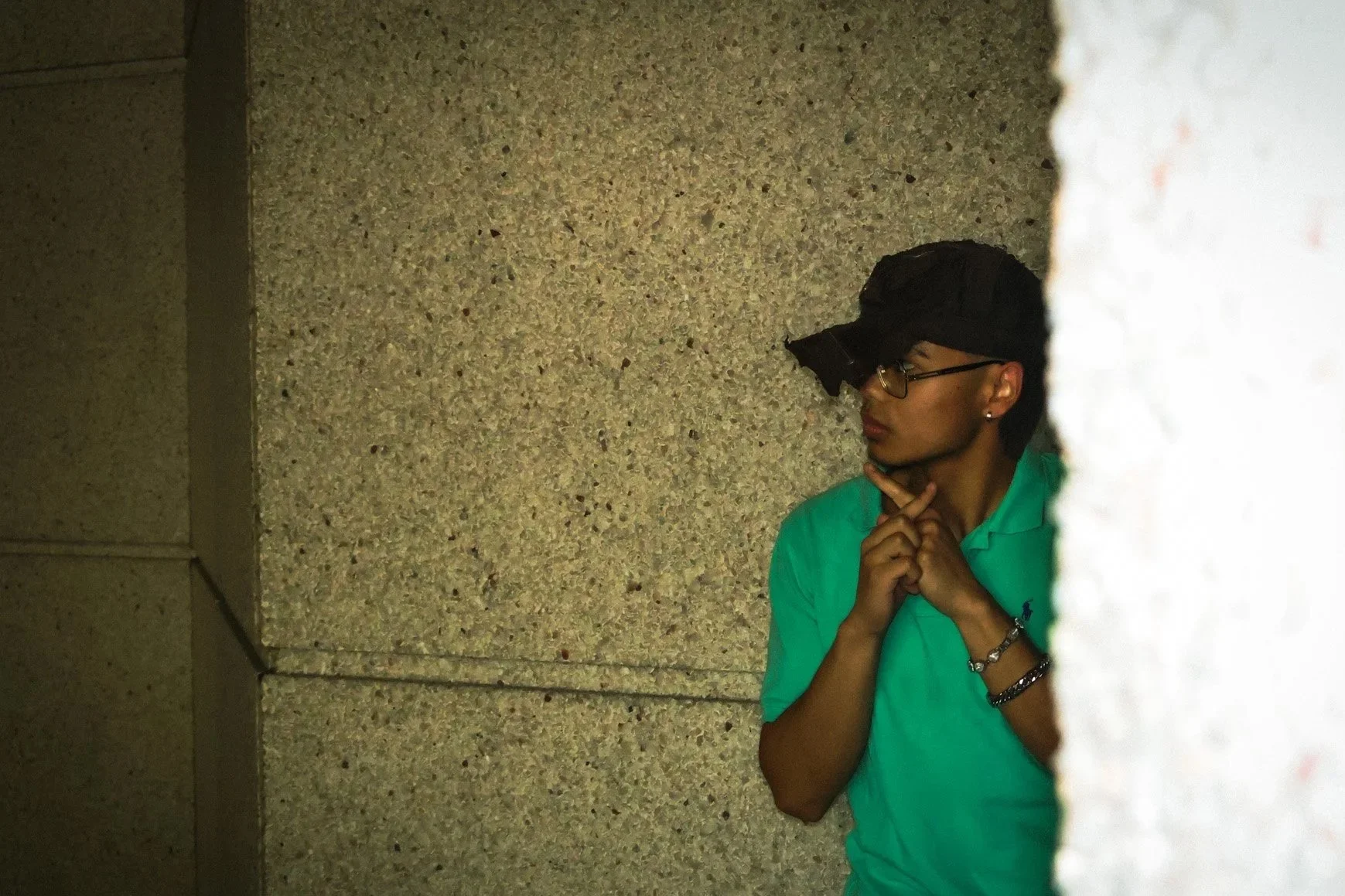 Young man wearing glasses and a turquoise polo shirt, posing with his hands under his chin, seen through a small gap in a concrete wall.
