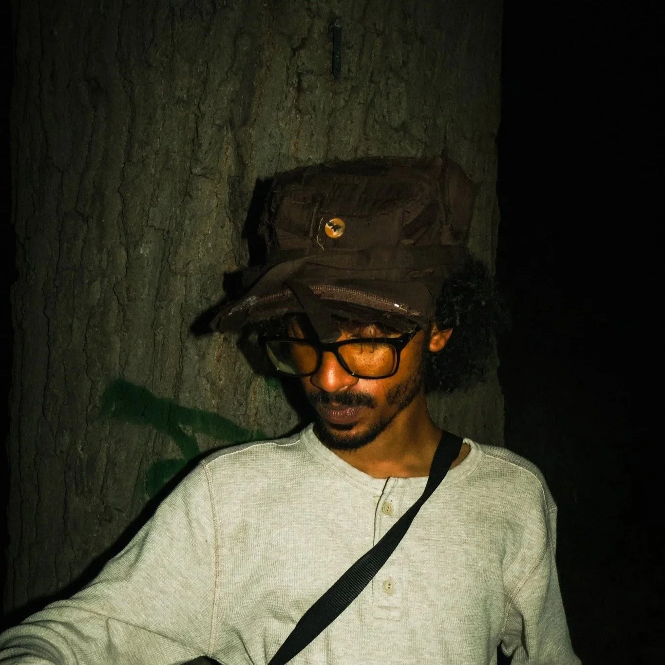 A man with glasses and dark curly hair standing against a tree at night. He is wearing a light-colored shirt and a backpack, with the bag's strap across his chest. The background is dark, indicating a nighttime setting.