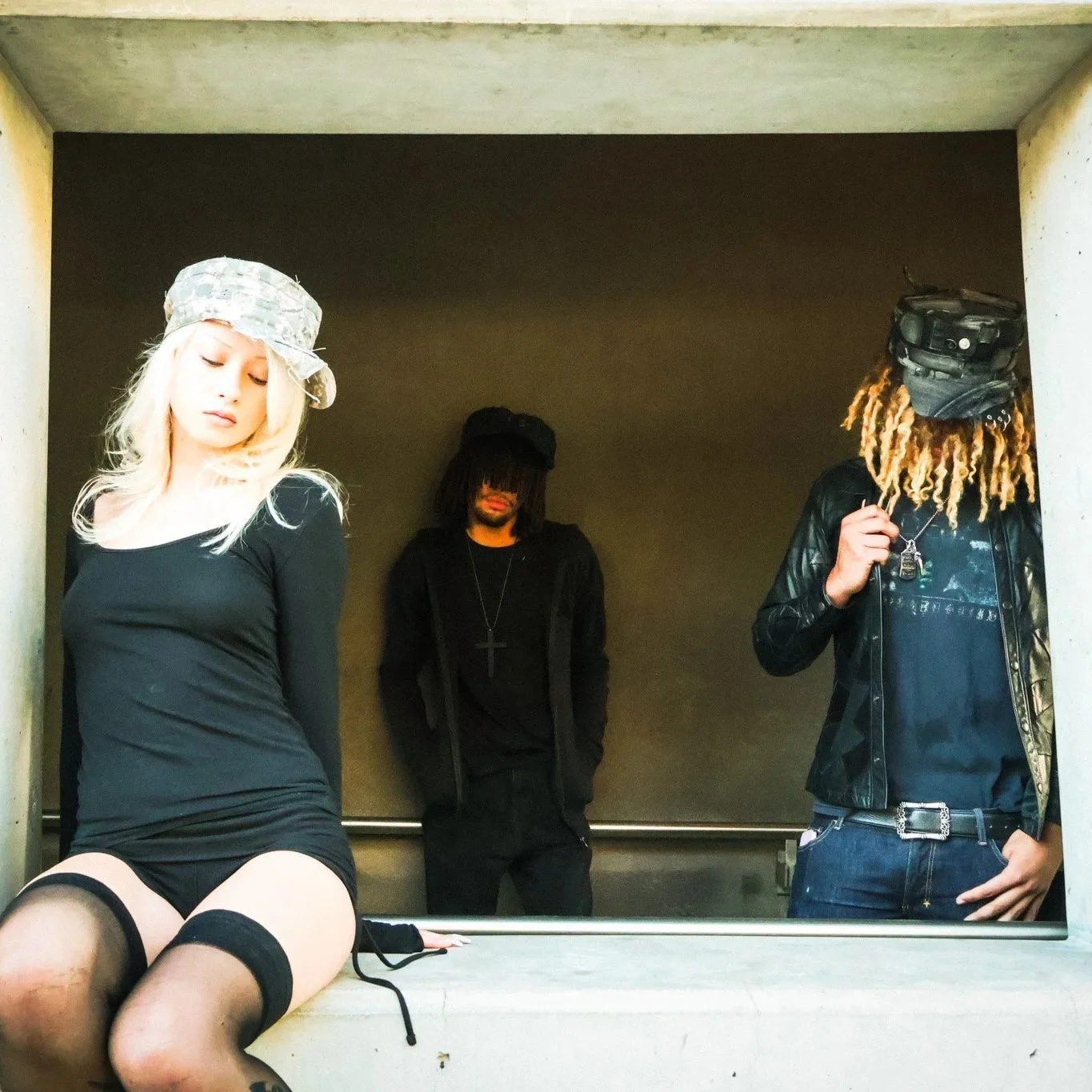 Three young adults in an indoor setting with a dark background. The woman on the left has long blonde hair, wears a black dress, and a camouflage hat. The man in the middle has dreadlocks, wears dark clothing, a cross necklace, and has his head sligh
