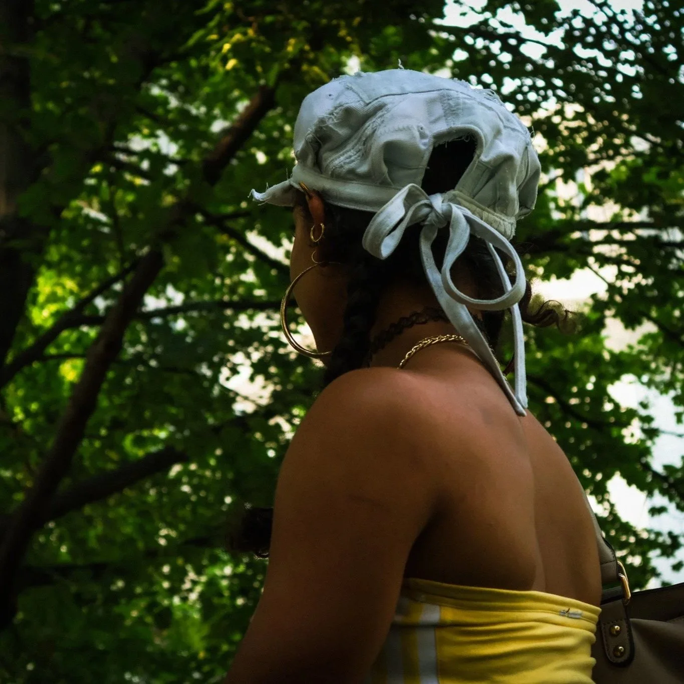 A woman with dark hair and gold jewelry wearing a white headscarf, gold ring, and chain, standing outdoors among green trees, with her back partially visible.