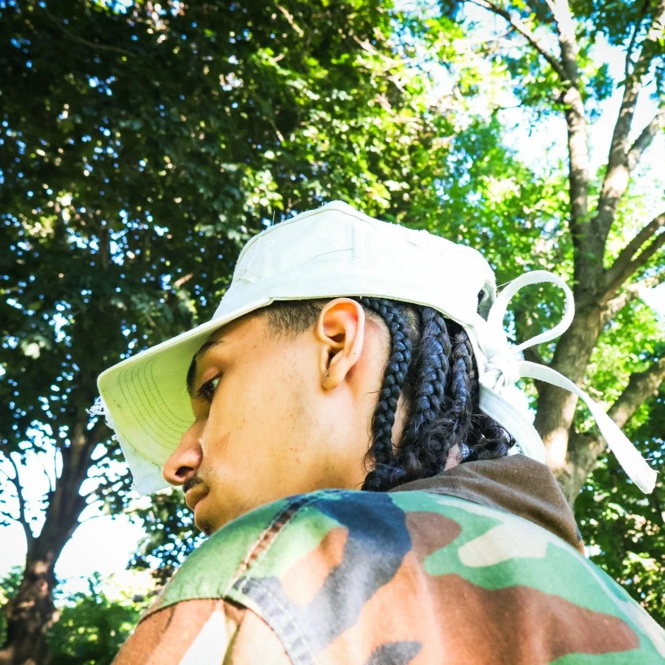 Side view of a person wearing a camouflage jacket and a white bucket hat with a leaf and tree background.