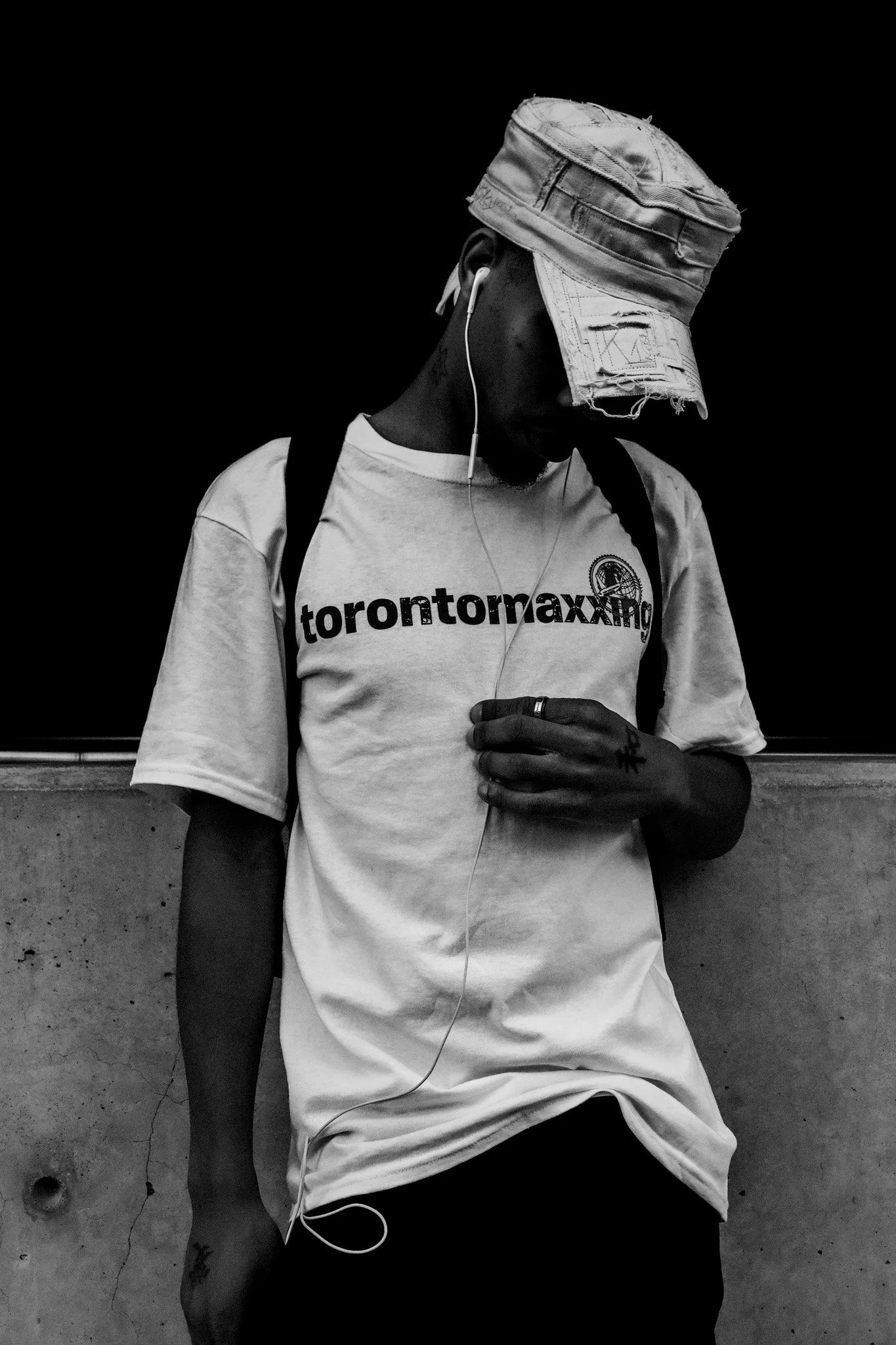A black and white photo of a person with dark skin wearing a distressed bucket hat, a Toronto maximum t-shirt, and earphones, leaning against a wall with arms resting on their knees, looking down.