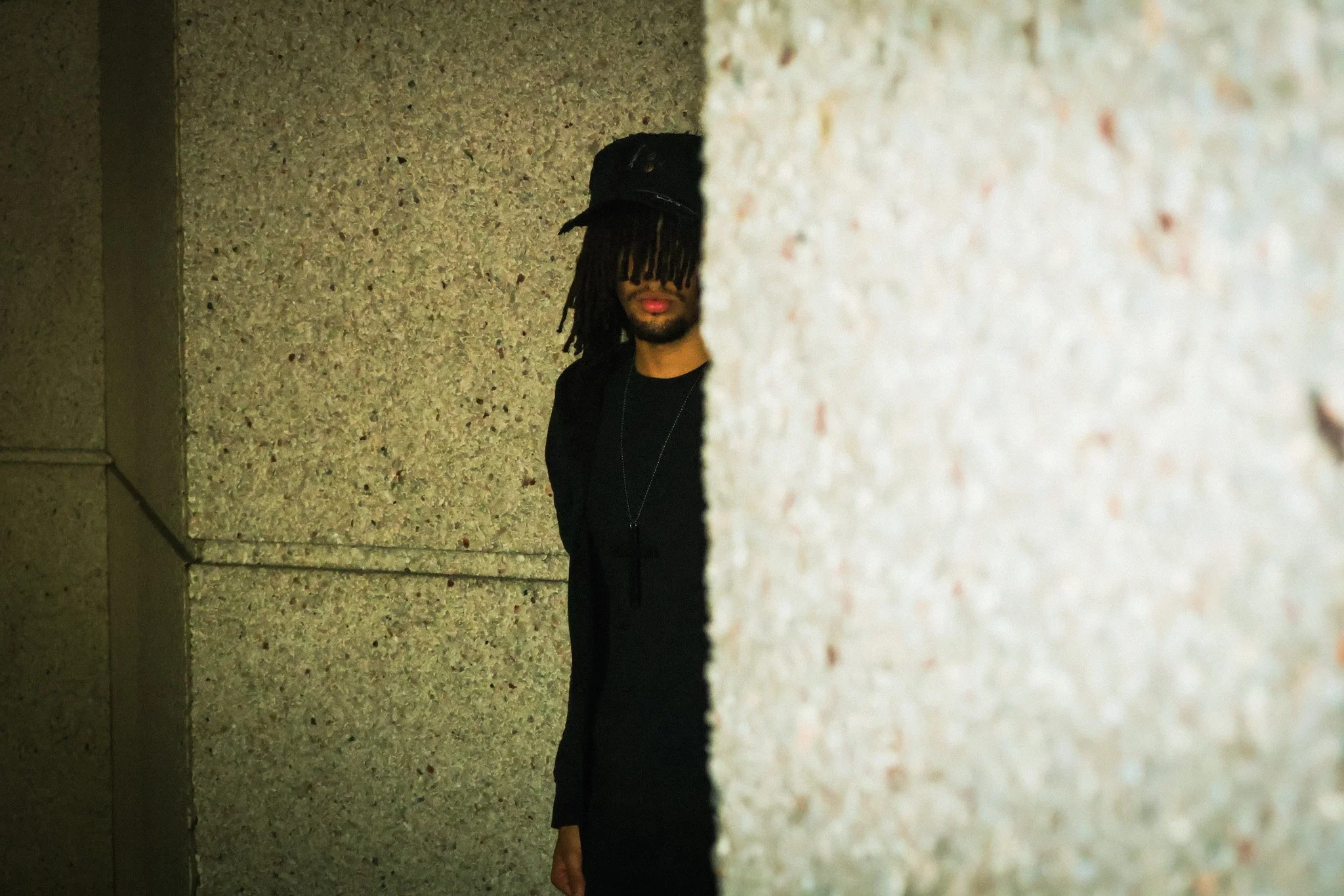 A person with dreadlocks covering part of their face, wearing a dark shirt, standing between two walls, one in front and one on the left.