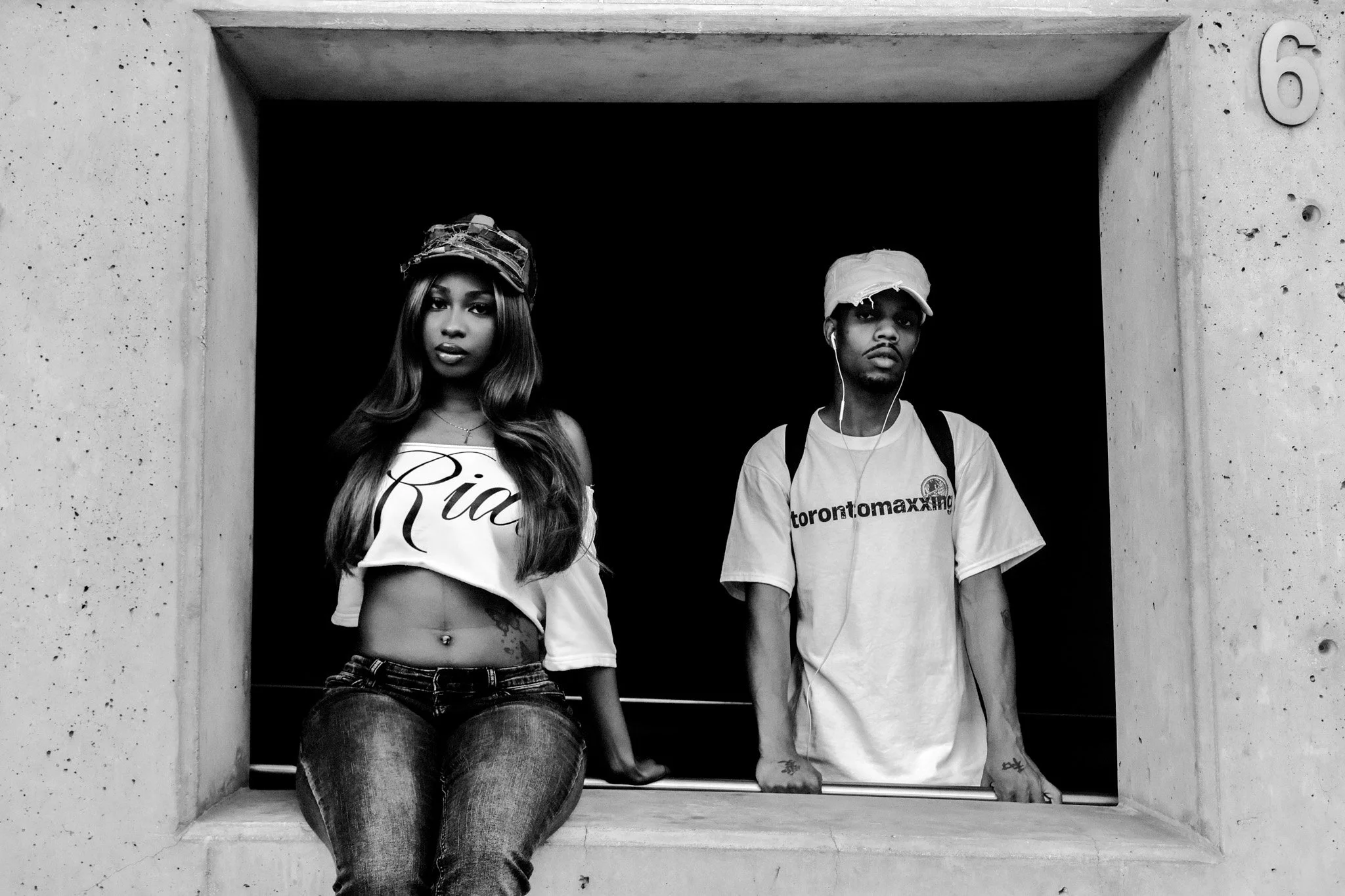 A black and white photo of a young woman sitting in a concrete window ledge, wearing a cropped top with 'Rie' printed on it, and jeans, with a hat and long hair. Next to her, a young man stands with earbuds, a backpack, a cap, and a T-shirt with some text, looking directly at the camera.
