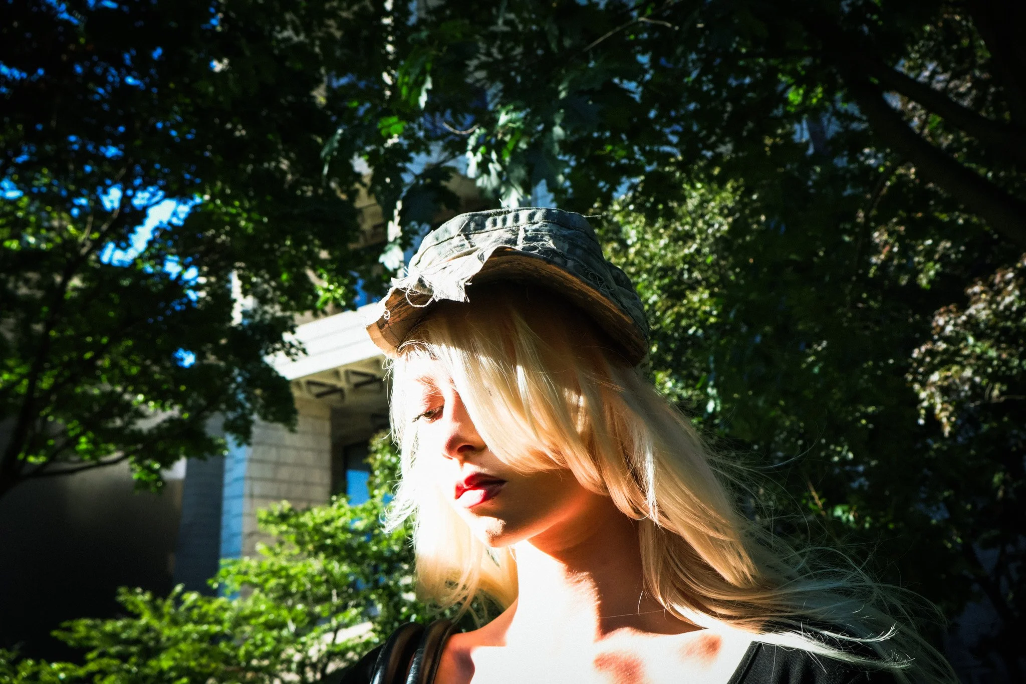 A woman with long blonde hair wearing a camouflage bucket hat and a black top, standing outdoors with green trees and a building in the background, looking down with sunlight on her face.