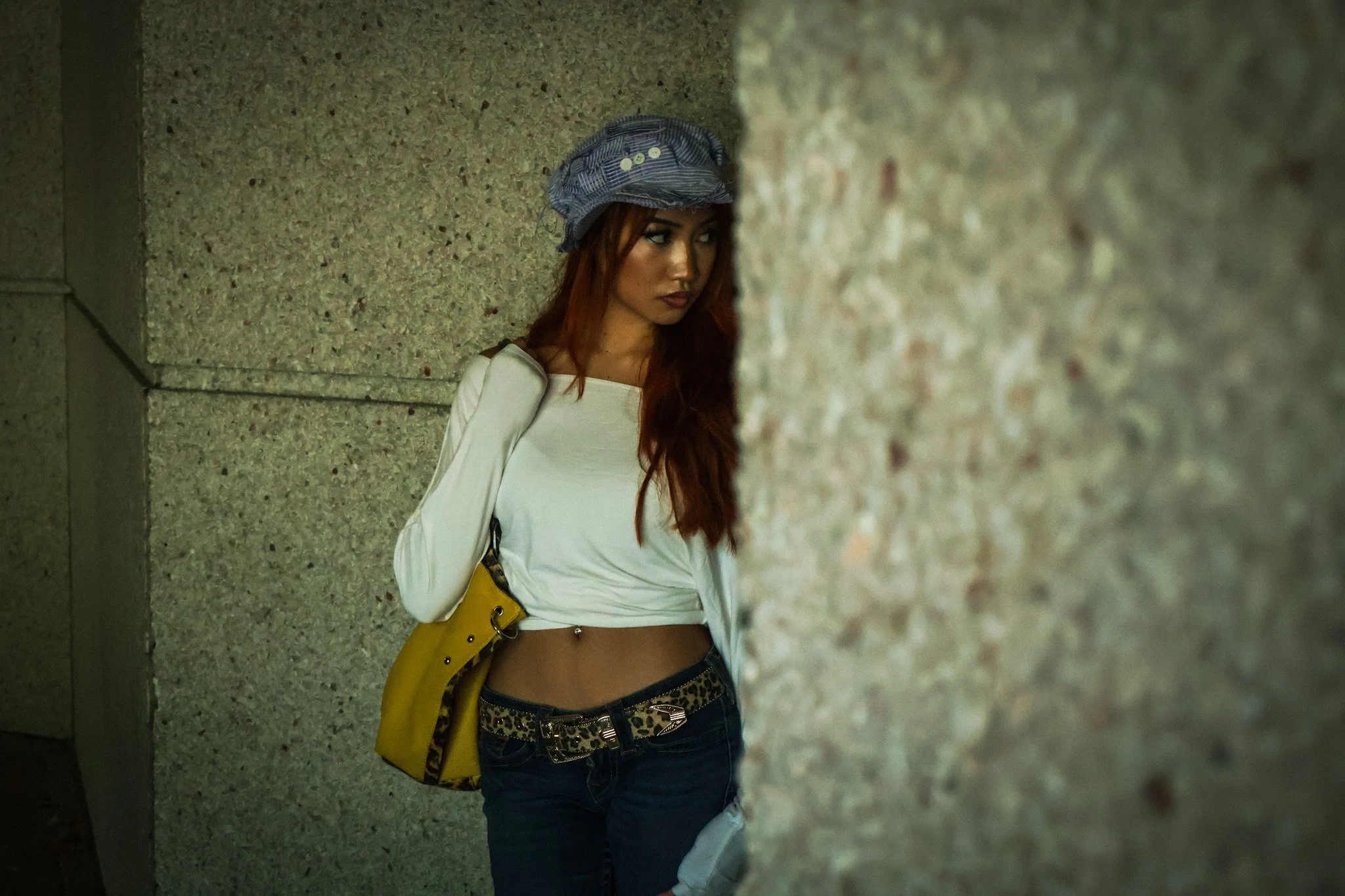 A young woman with long red hair, wearing a blue striped hat and a white long-sleeve shirt, partially tucked into dark pants with a leopard print belt, stands against a textured stone wall, looking to her right with a serious expression.