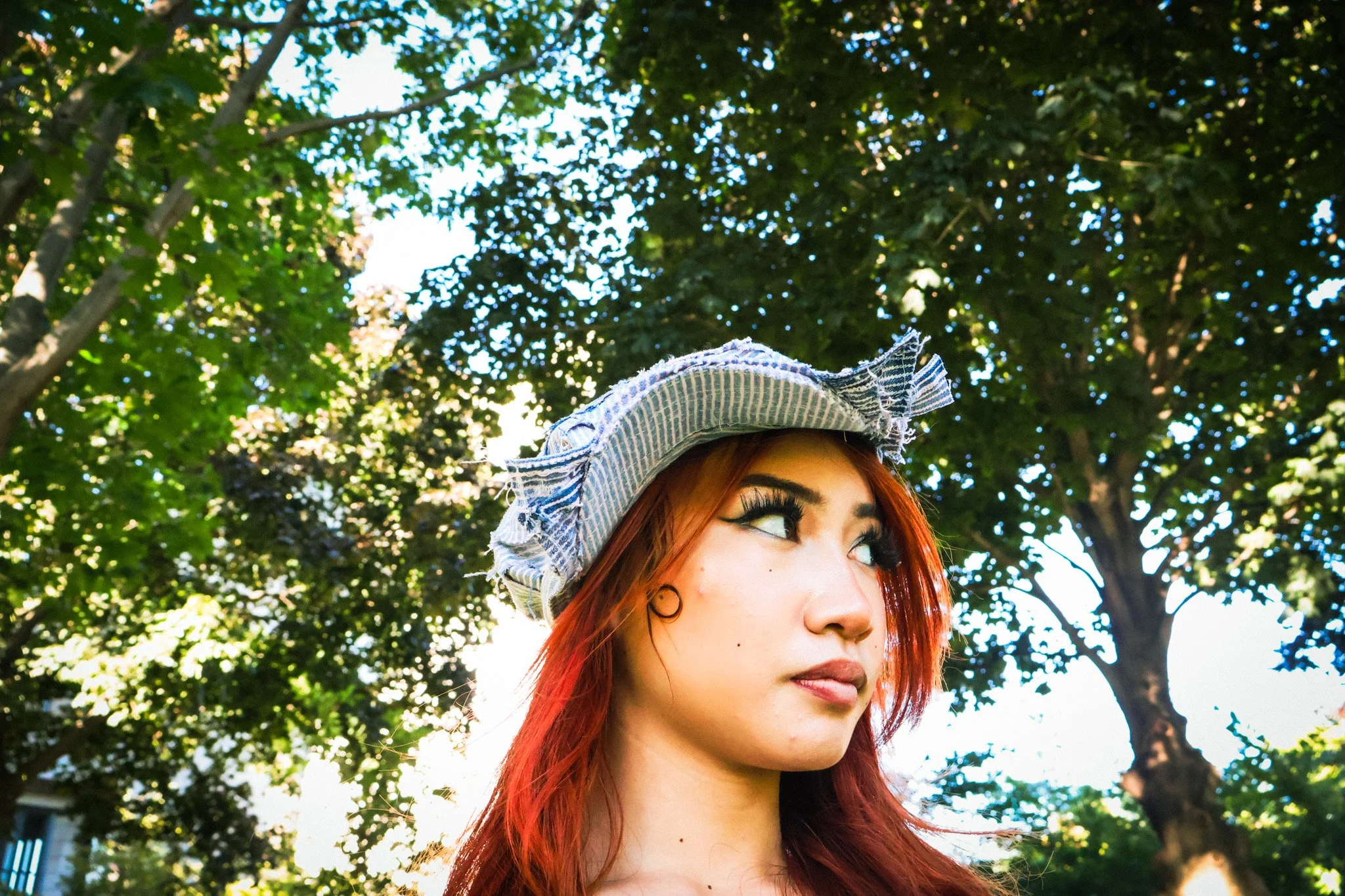 A woman with red hair wearing a striped sun hat and makeup, looking to the side outdoors in a park with green trees and sunlight.