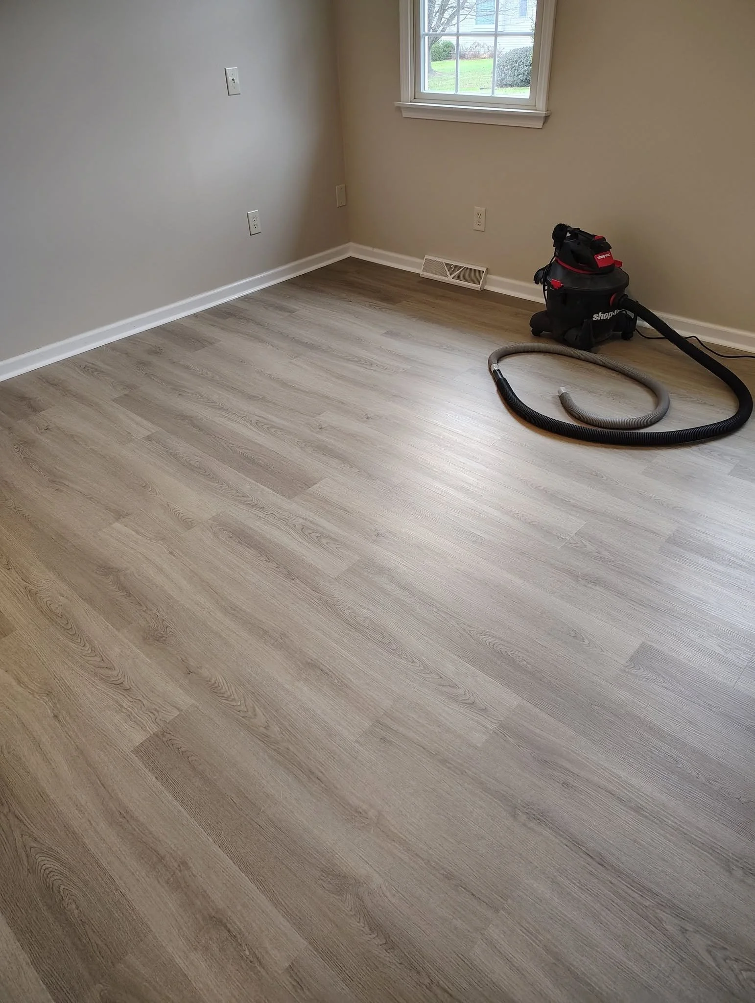An empty room with light wood flooring, a window showing a lawn and bushes outside, and a vacuum cleaner with a hose on the floor in the corner.