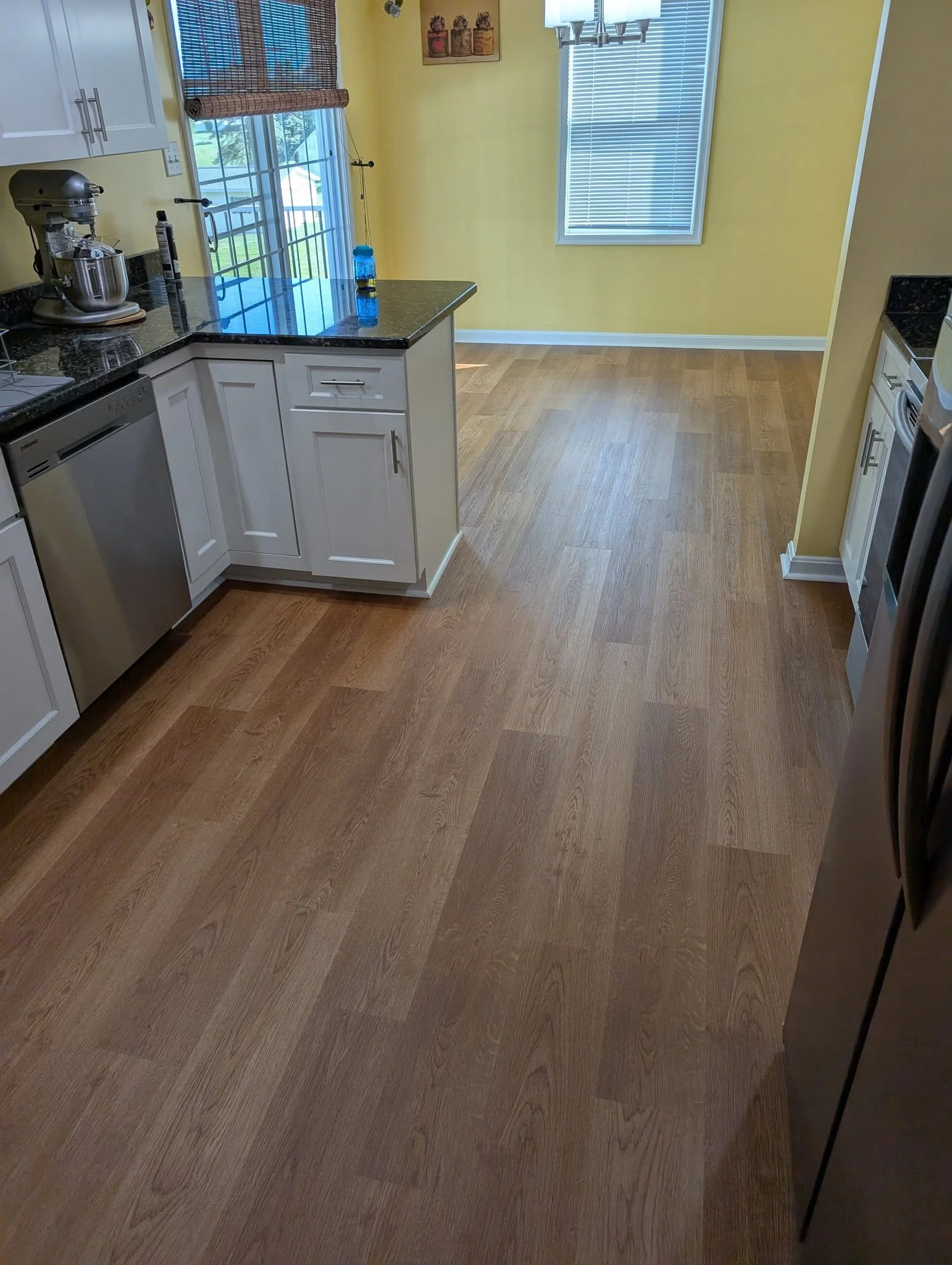 Kitchen with light yellow walls, white cabinets, black granite countertops, a window with blinds, a chandelier, and hardwood floors. There is a stand mixer and a water bottle on the counter.