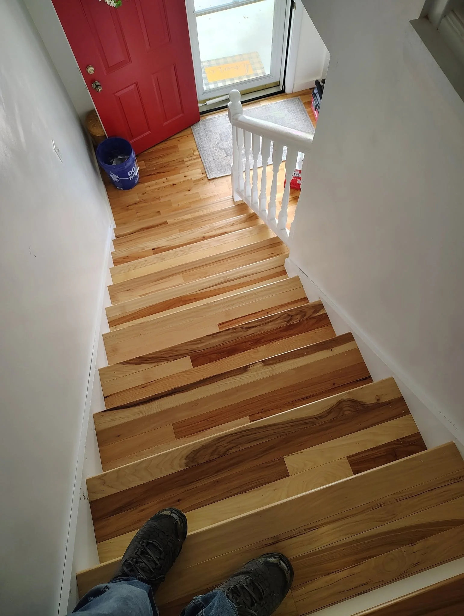 Photo taken from the top of a staircase looking down at wooden stairs, with a person's feet in hiking boots. At the bottom, there's a red front door with a white door frame, a gray doormat, a blue recycling bin, and a glass door leading outside. A ye