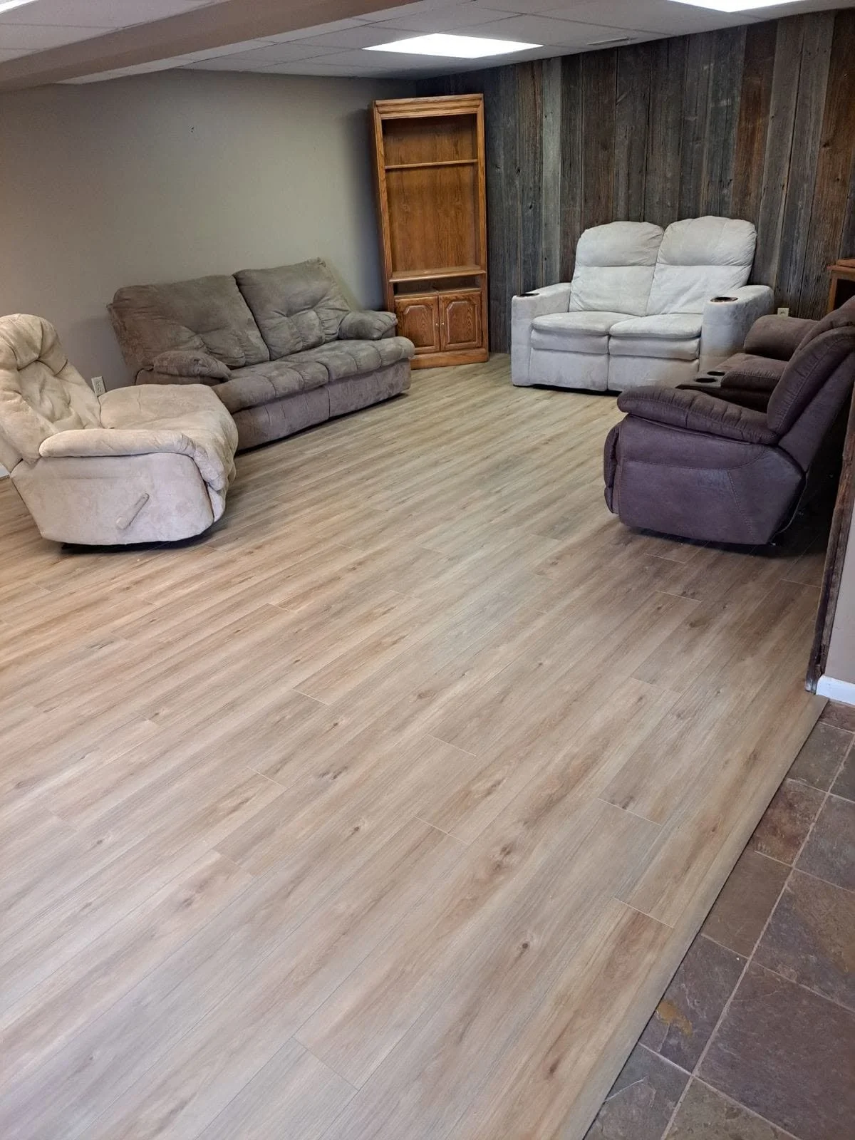 A living room with beige, brown, and white recliner sofas and a wooden bookshelf against a wood-paneled wall.