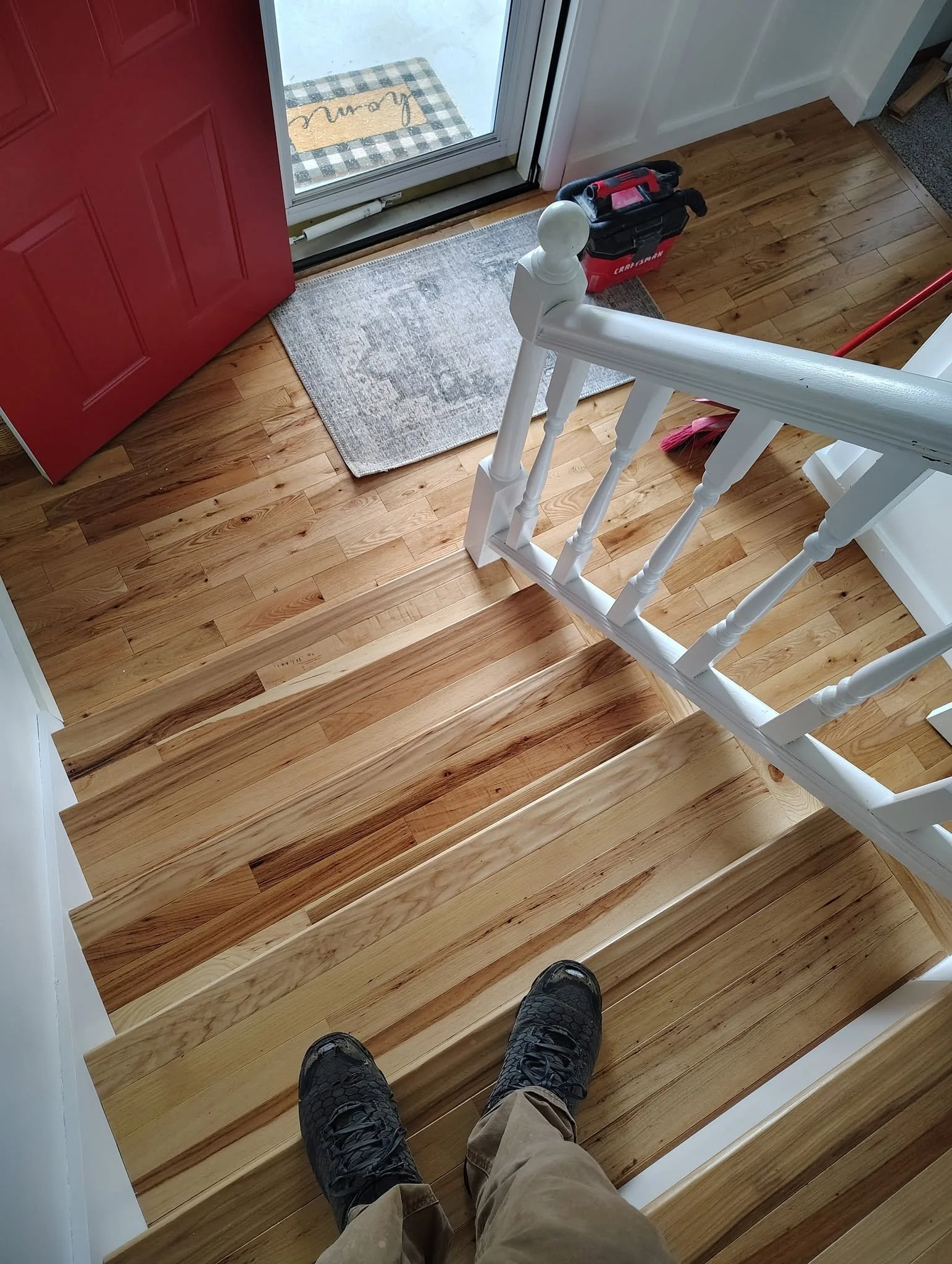 View of a person standing on a staircase holding a camera or phone, showing black shoes, beige pants, and the staircase with wooden steps and a white railing. The perspective looks downward from the top of the stairs.
