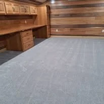 Empty room with wooden panel walls, built-in wooden desk with drawers, and a light-colored carpeted floor.