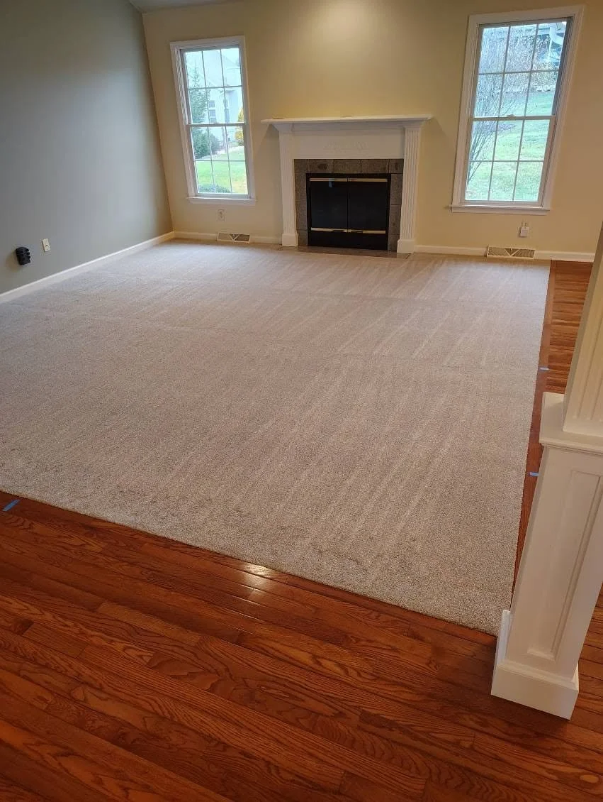 Empty living room with beige walls, hardwood flooring, a fireplace with a white mantel, two windows, and a large beige carpet.
