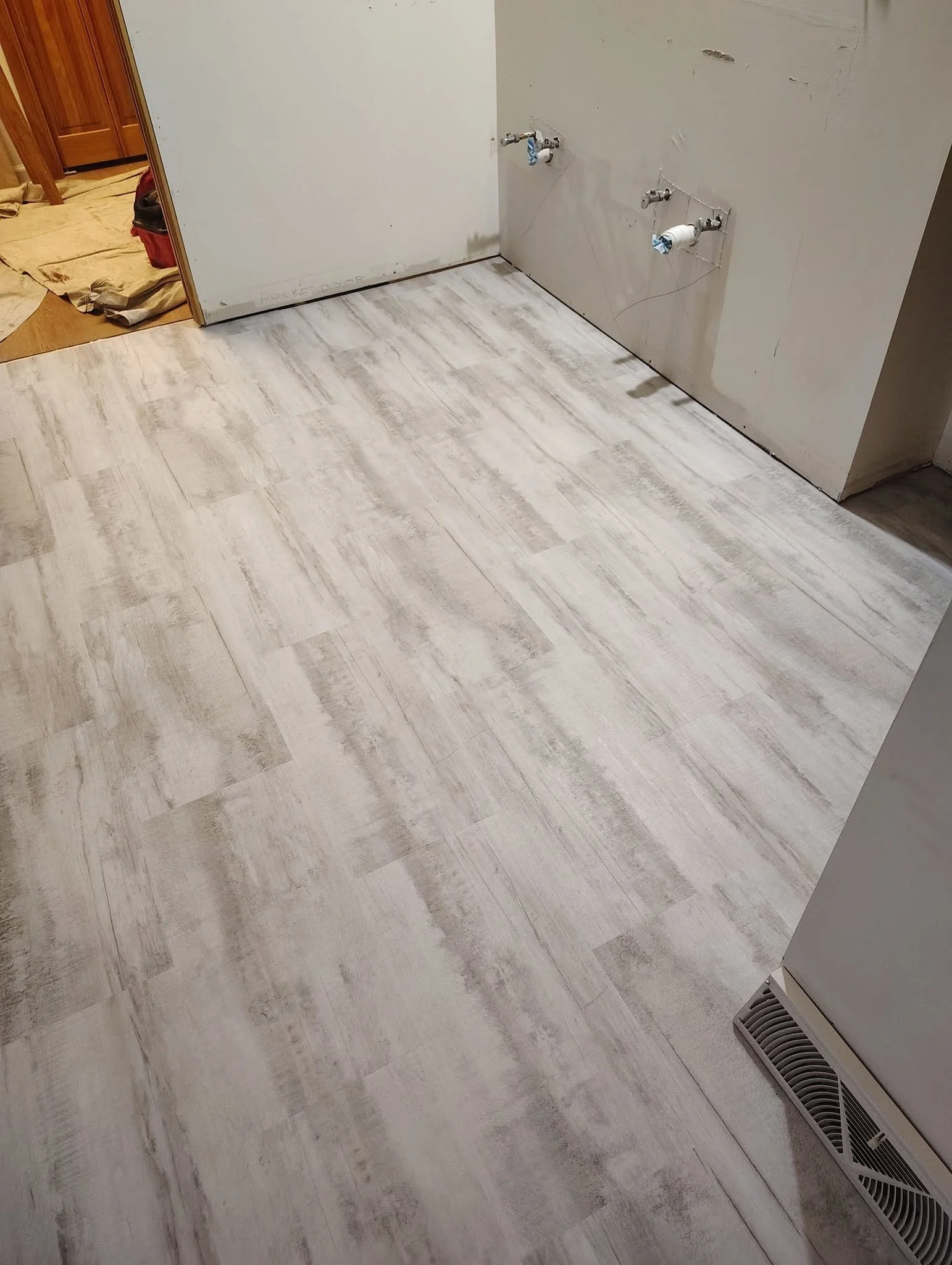 A room under renovation with a white wood-look floor, a section of unfinished wall with plumbing fixtures, and a vent near the floor.