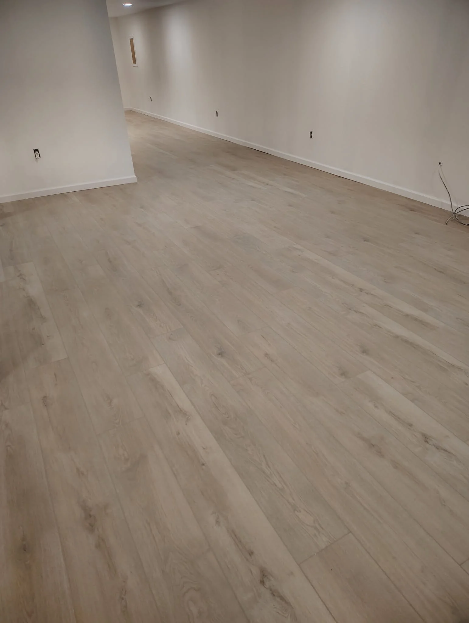 Empty room with white walls, light-colored wood flooring, and electrical outlets. Some wires are visible on the floor near the wall.