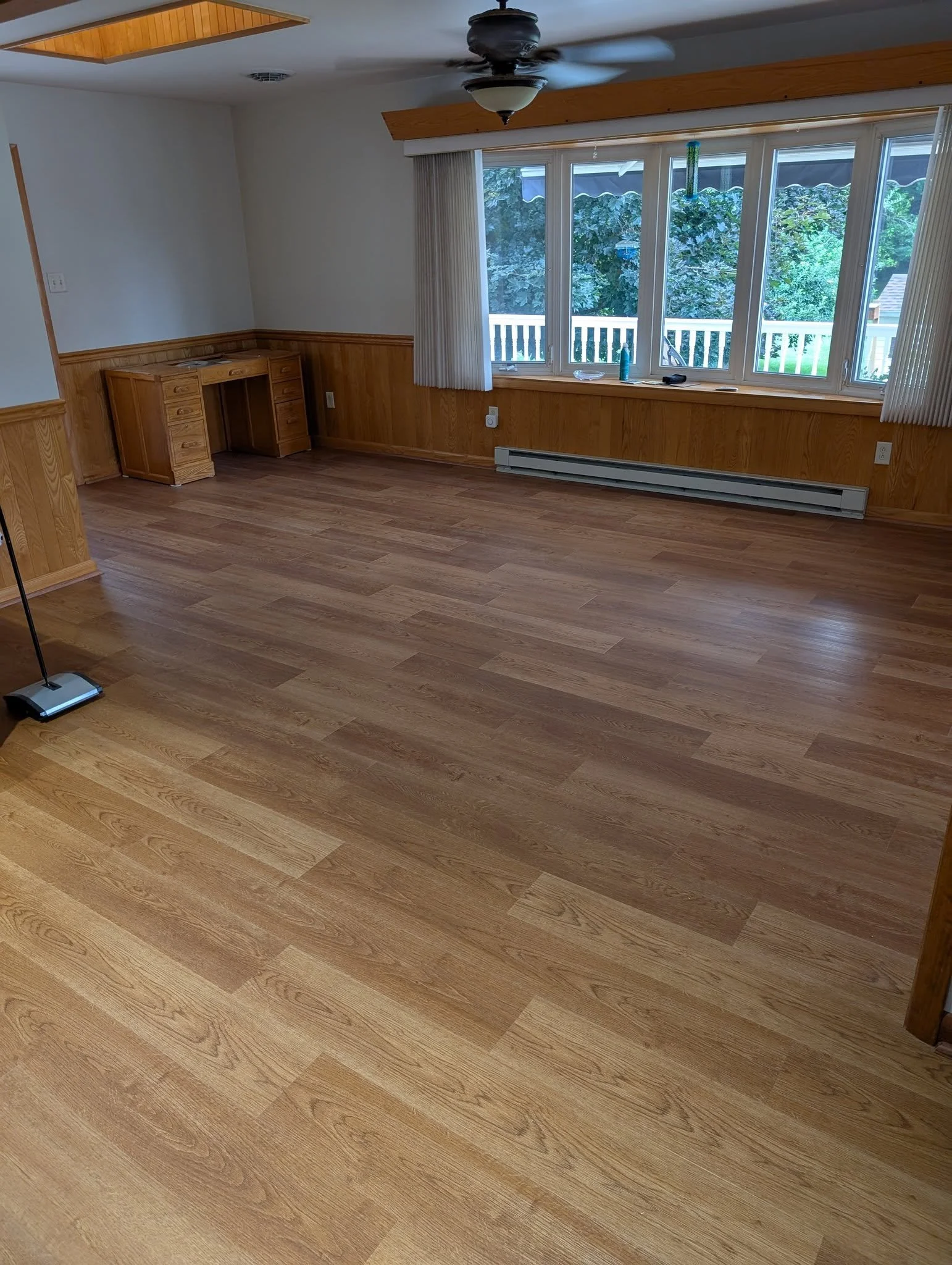 Empty living room with hardwood floors, a large window with vertical blinds, a ceiling fan, and a small wooden desk in the corner.