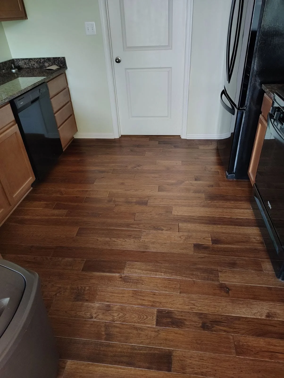 Kitchen with wooden flooring, partially visible black dishwasher, white door, brown cabinets, and black refrigerator.