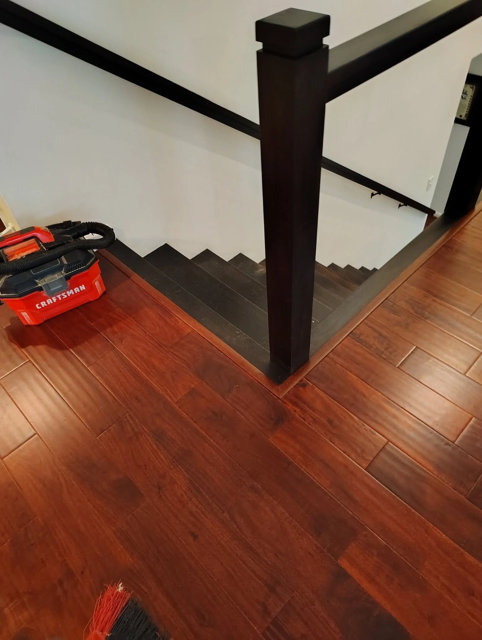 View of a staircase with wooden steps, black handrail, and white walls, seen from above, with part of a red and black vacuum cleaner in the lower left corner.