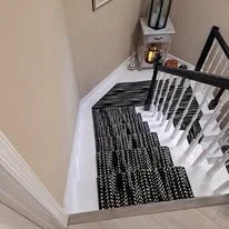 Staircase with black and white striped rug, beige walls, wooden handrail, and a small trash can at the top of the stairs.