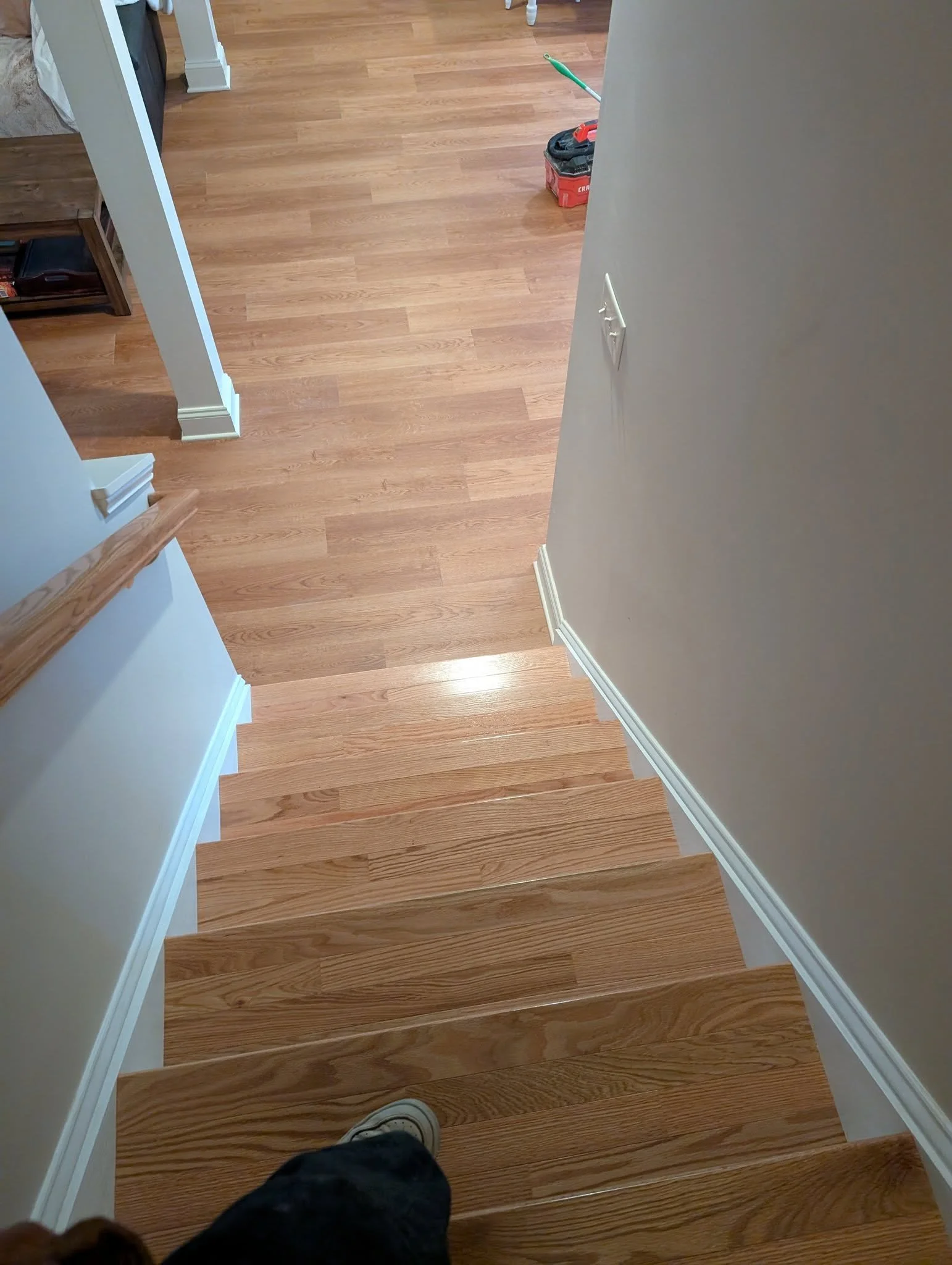 View of a hardwood staircase from the top, with a person's foot visible at the bottom. Part of a room with a wooden shelf and a door entrance can be seen, and a toolbox is on the floor.